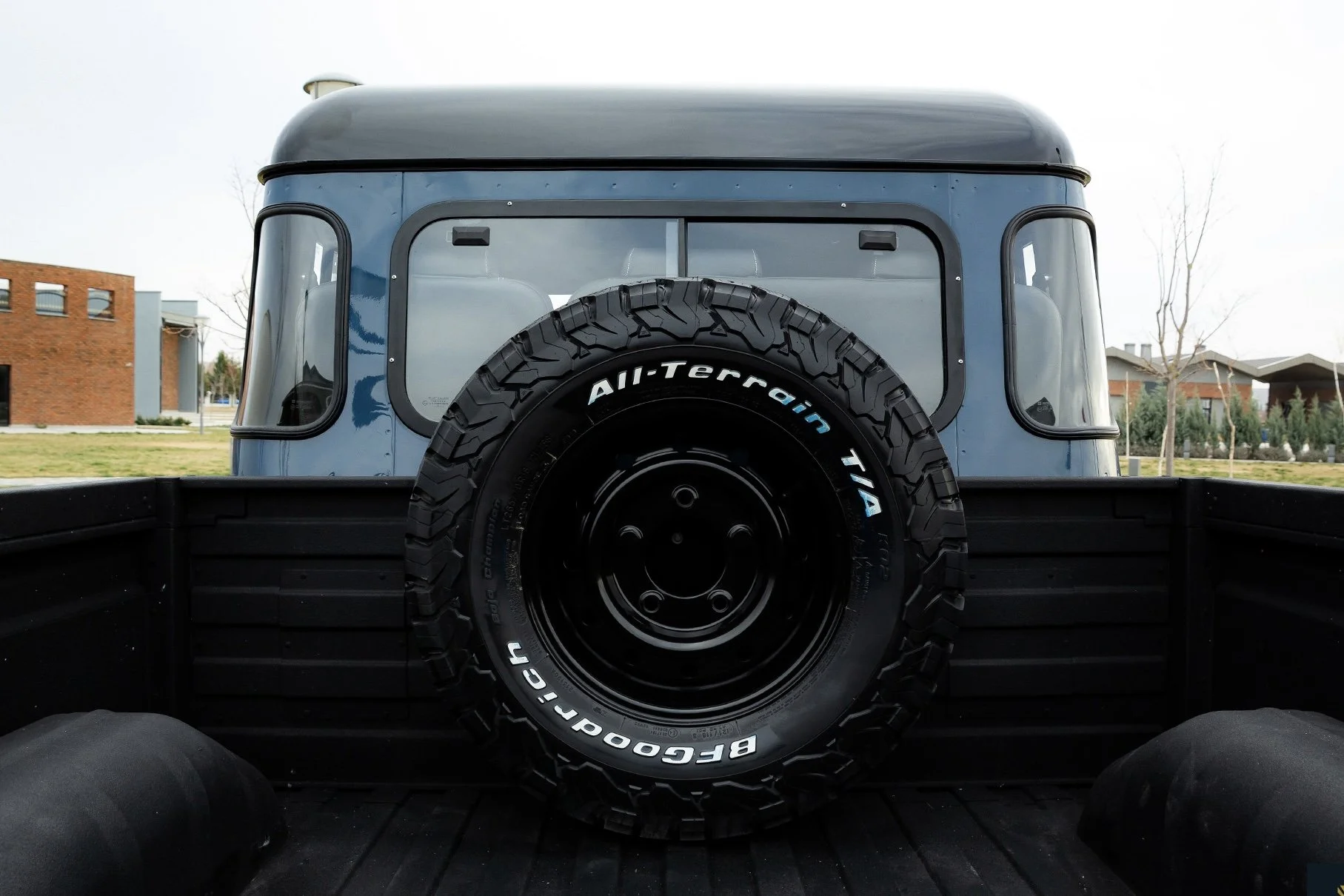 View of the back of a truck bed with a large off-road tire mounted in the center, featuring a blue and black color scheme and the words 'All-Terrain T/A' and 'BFGoodrich' on the tire.