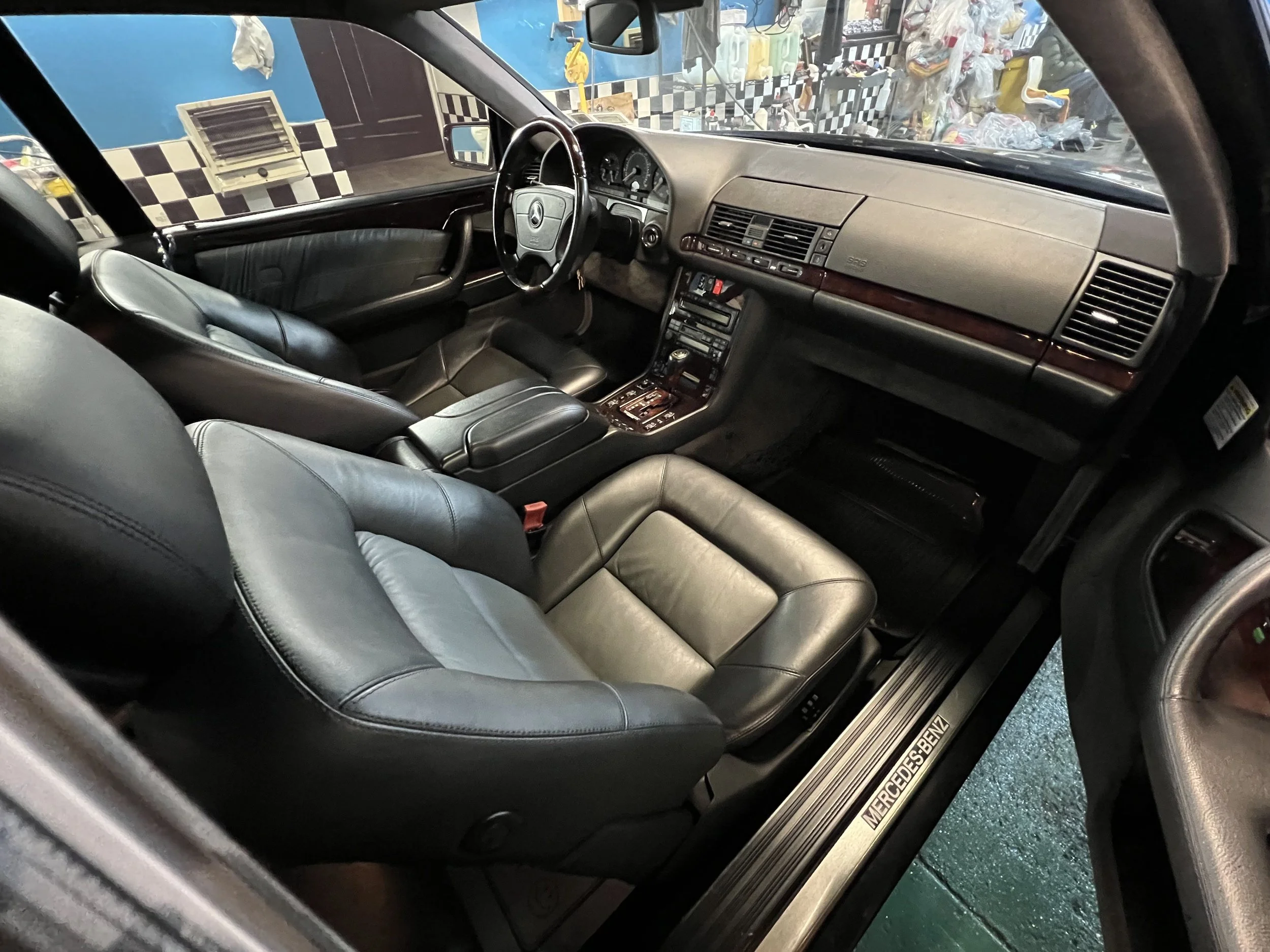 The image shows the interior of a classic Mercedes-Benz car with black leather seats, a dashboard with wood trim, and a steering wheel with the Mercedes-Benz logo. The front seats and part of the door panel are visible.