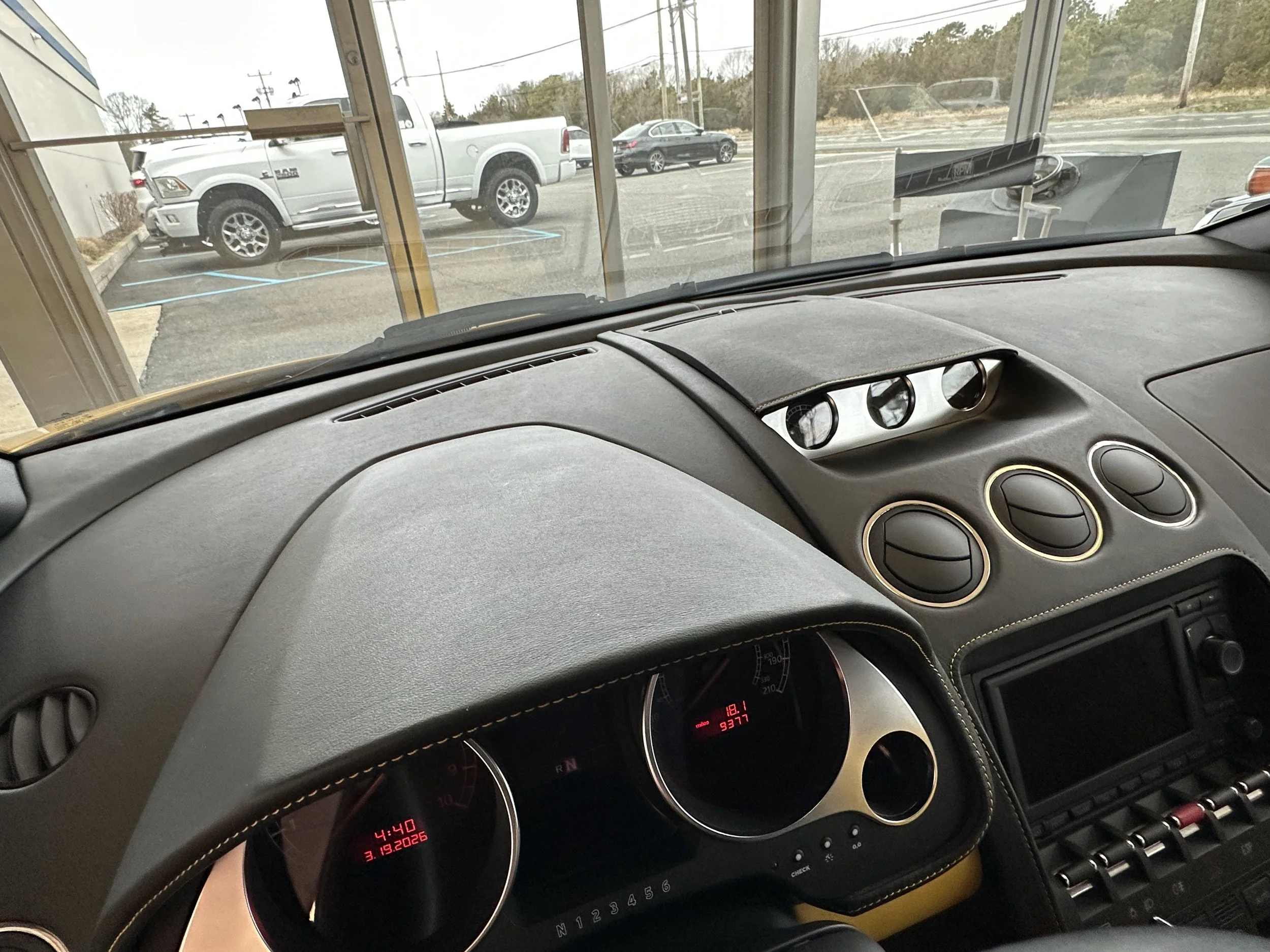 View from inside a vehicle's dashboard looking out through a large front window to a parking lot with a silver pickup truck and other cars, with a cloudy sky and trees in the background.