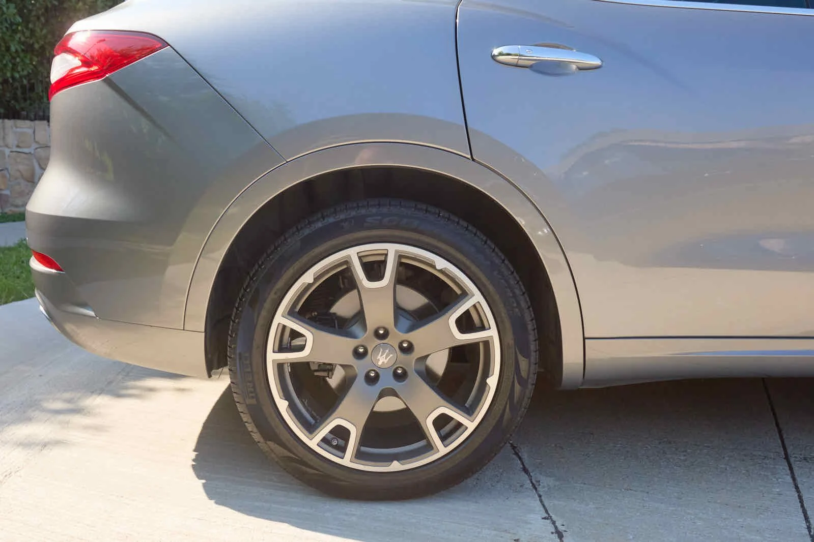 Rear side view of a gray luxury sedan car showing the rear wheel and part of the car body, parked on a concrete driveway during daytime.