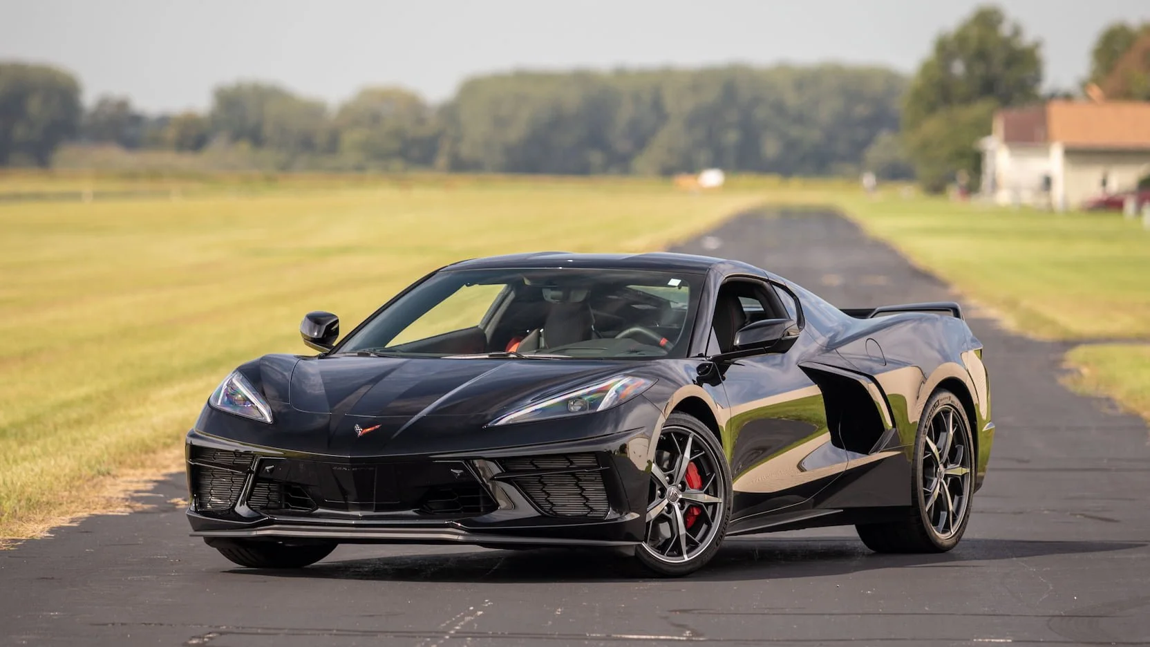 Black sports car parked on a paved road in a rural area with fields and some houses in the background.