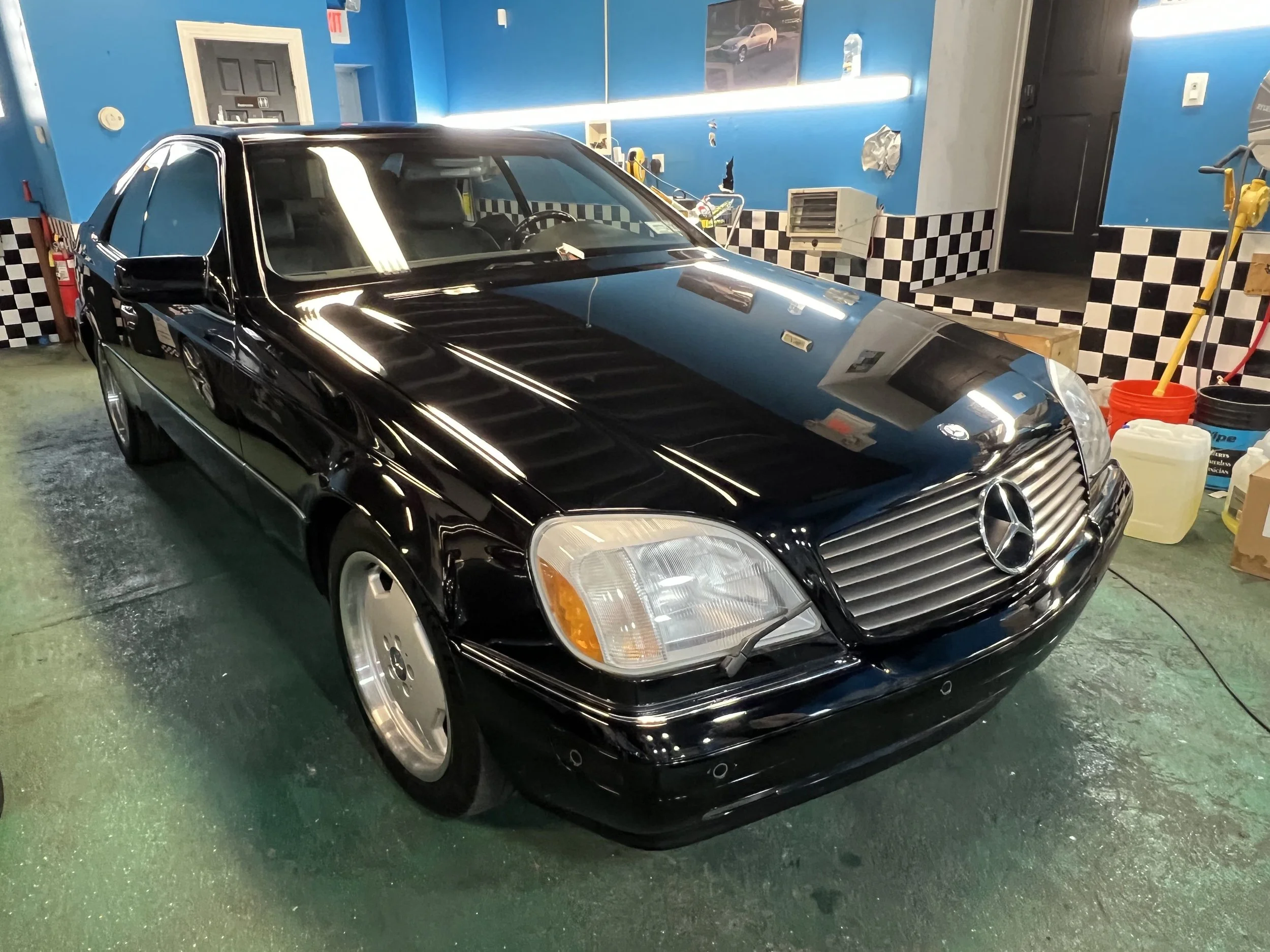 Black Mercedes-Benz car inside a garage with blue walls, checkered black and white tiles, and cleaning supplies on the right side.
