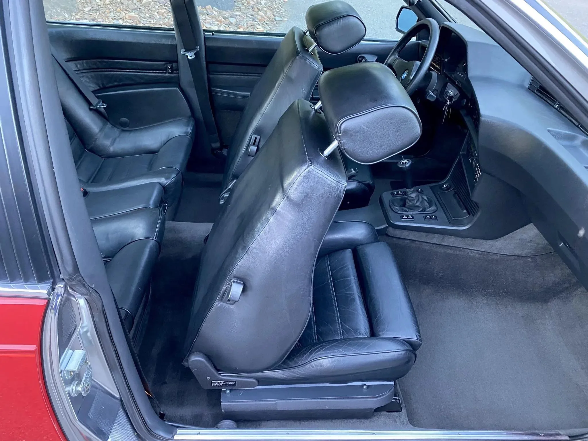 The interior of a vintage car showing black leather seats, a manual gear shift, dashboard with controls, and a steering wheel. The front seats are one behind the other, with the back seats visible behind the front seats.