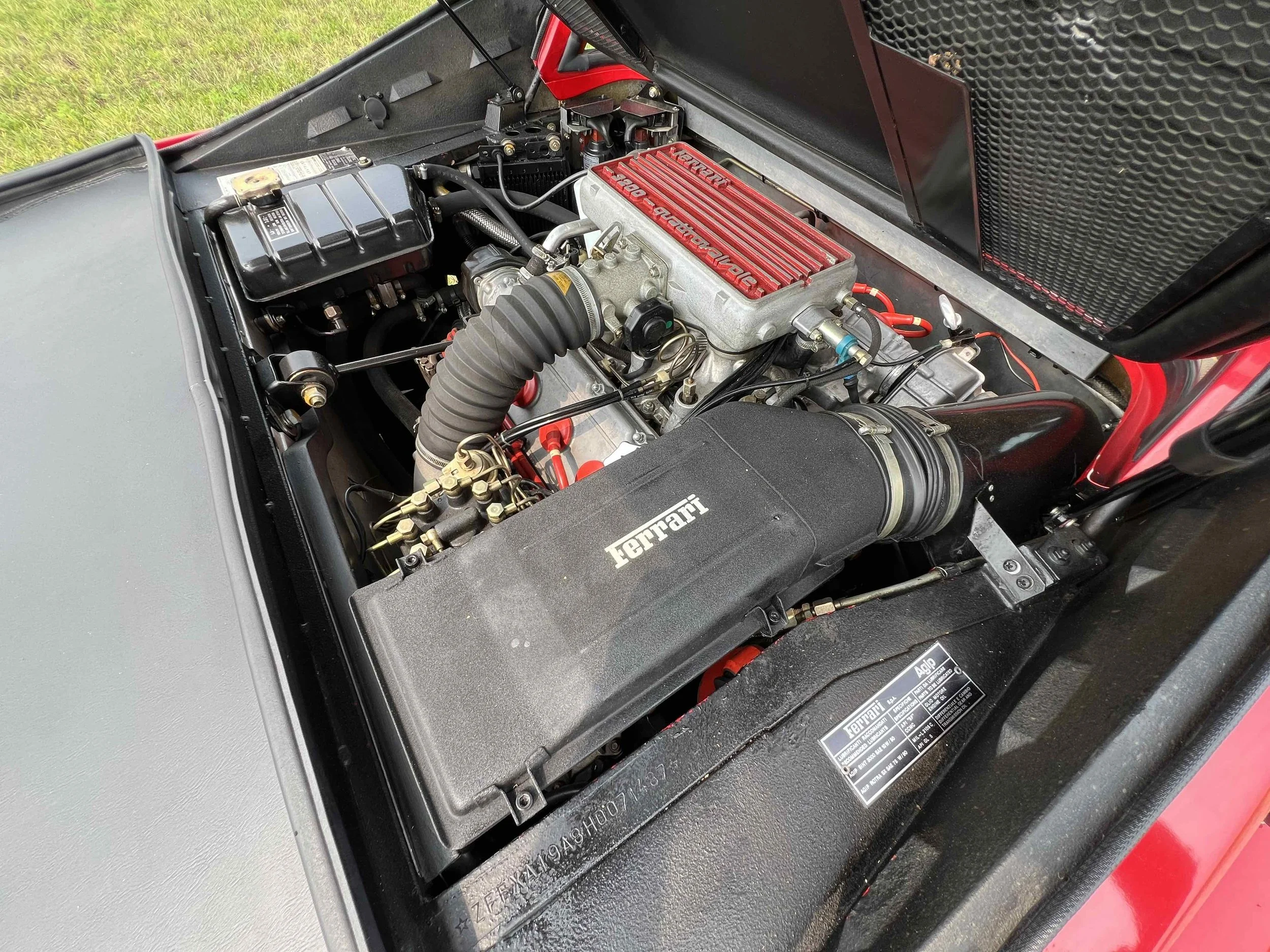 Close-up view of a Ferrari engine bay, showing a large air intake with the Ferrari logo, red engine valve covers, and various hoses and components.