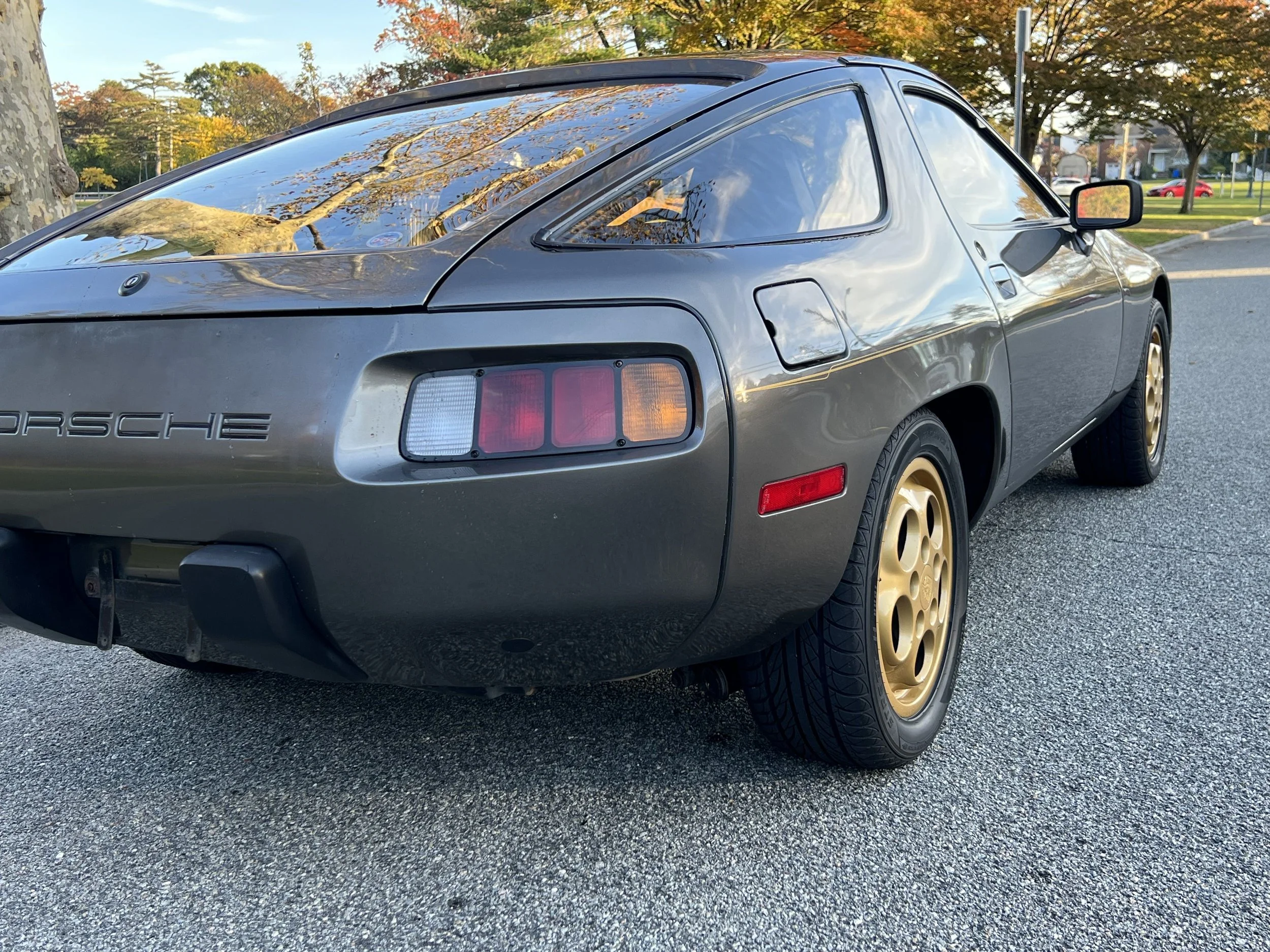 A gray vintage Porsche sports car with gold wheels parked on a street during autumn, reflecting trees in the rear window, with a park and trees with fall foliage in the background.