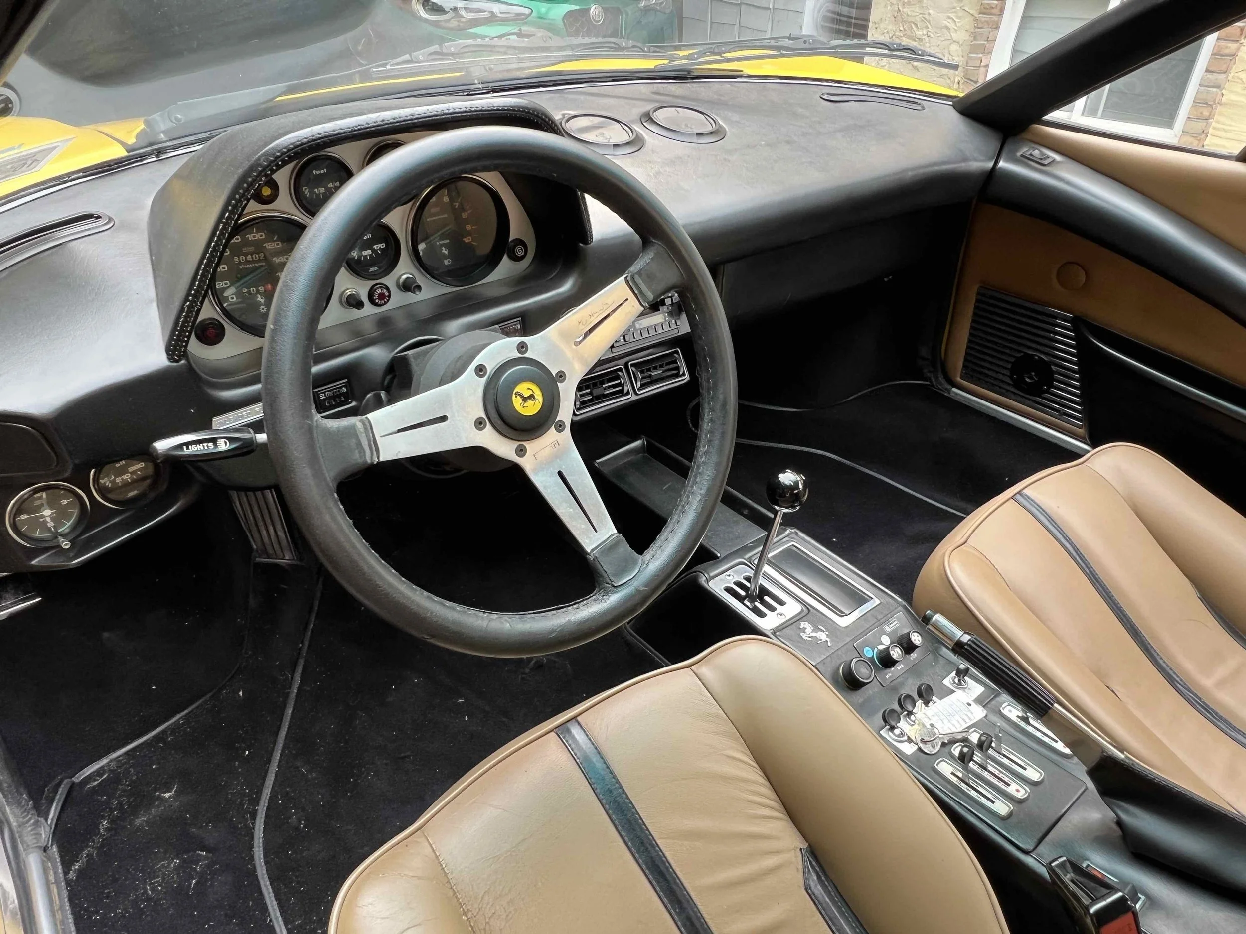 Interior of a classic Ferrari sports car, featuring a black leather steering wheel with a yellow Ferrari emblem, tan leather seats, a black dashboard with multiple gauges, and a central gear shifter.