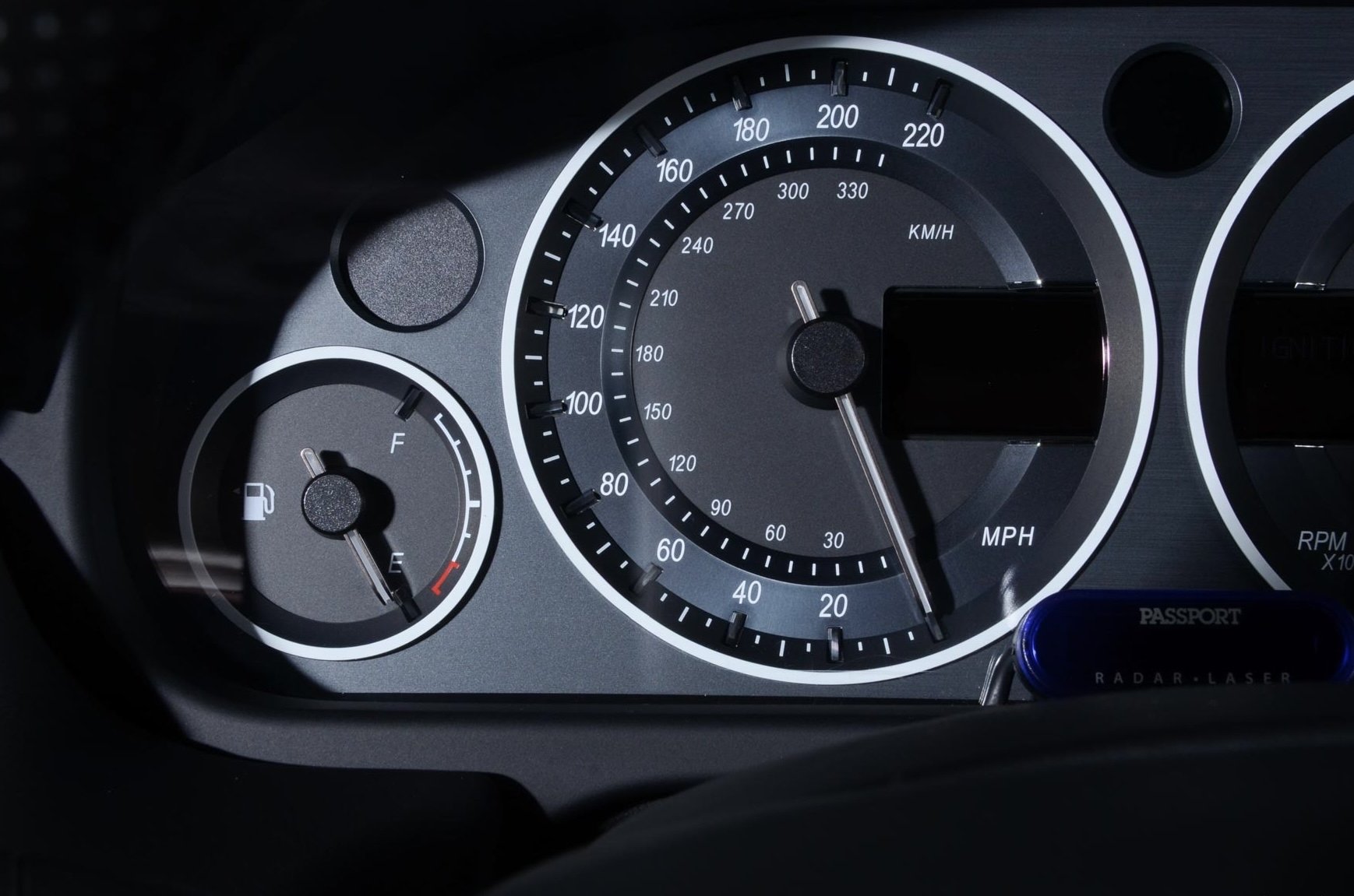 Close-up of a car dashboard showing the fuel gauge, speedometer, and a digital display, with a GPS device labeled 'PASS' attached to the lower right.
