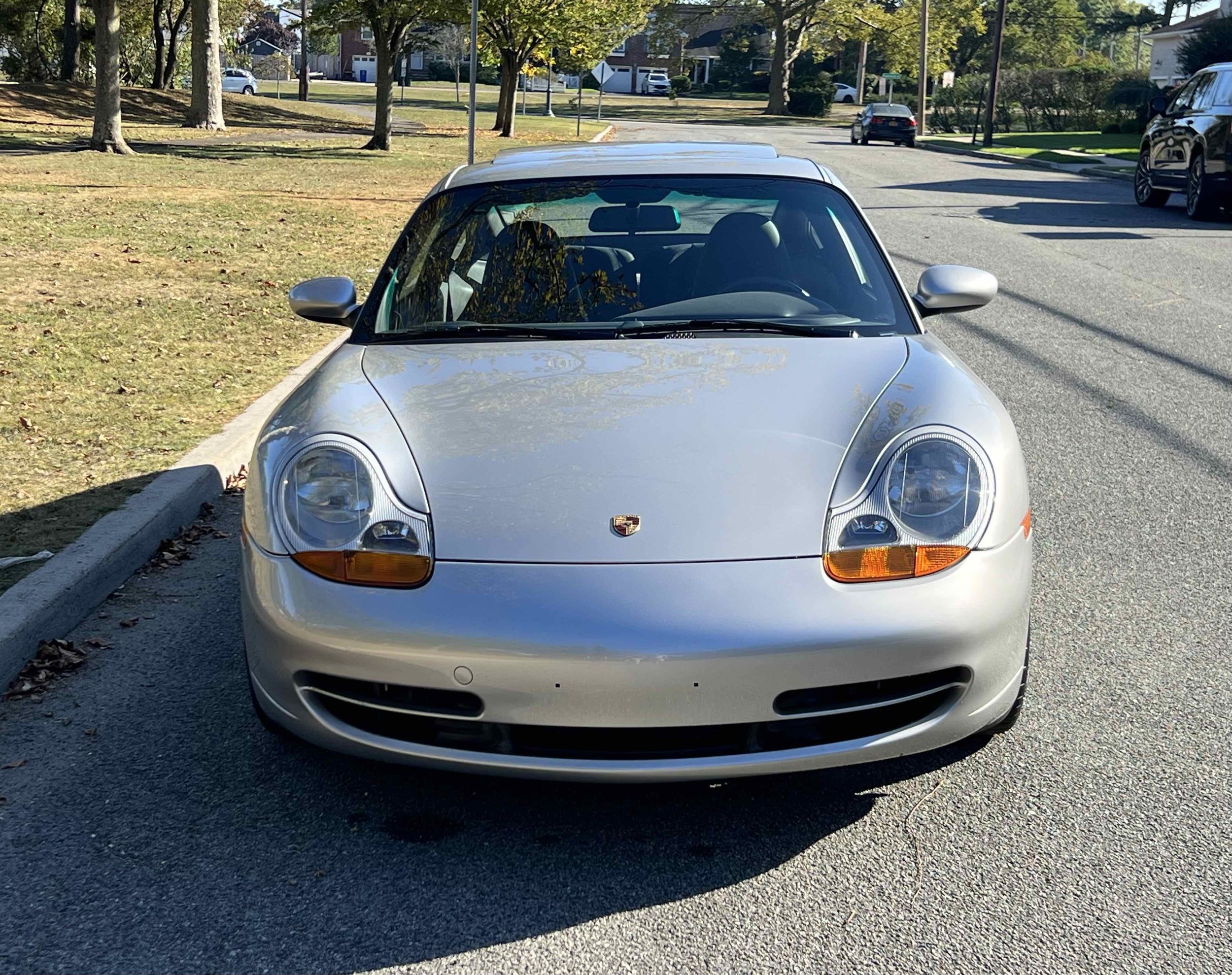 Front view of a silver Porsche sports car parked on a residential street with trees and other cars in the background.