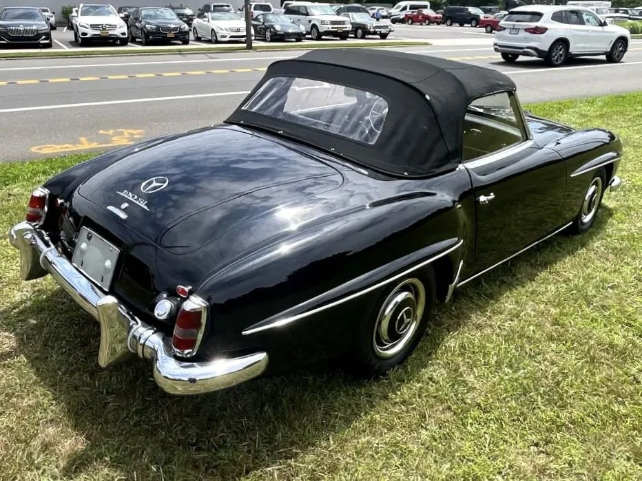 Black vintage Mercedes-Benz convertible car with a soft top, parked on a grassy area near a parking lot with several modern cars.