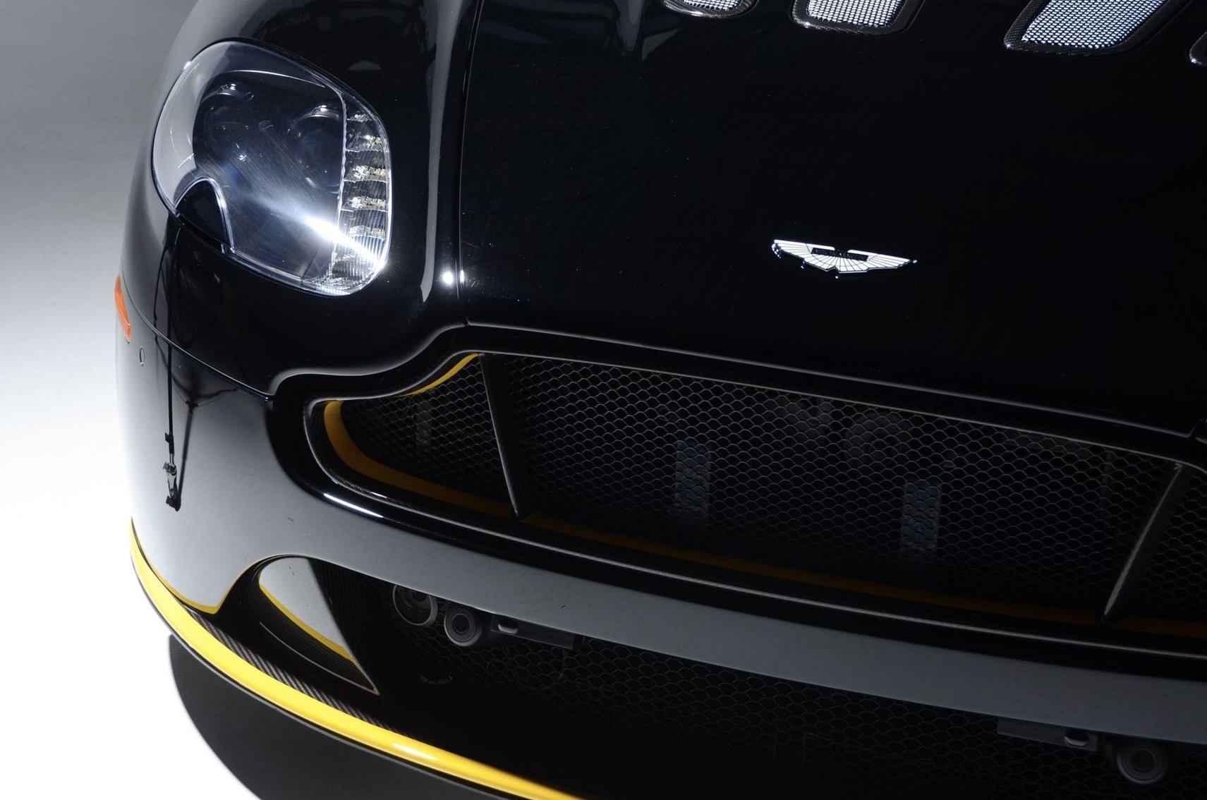 Close-up view of the front left corner of a black sports car, showing the headlight, front grille, and part of the yellow stripe on the bumper.