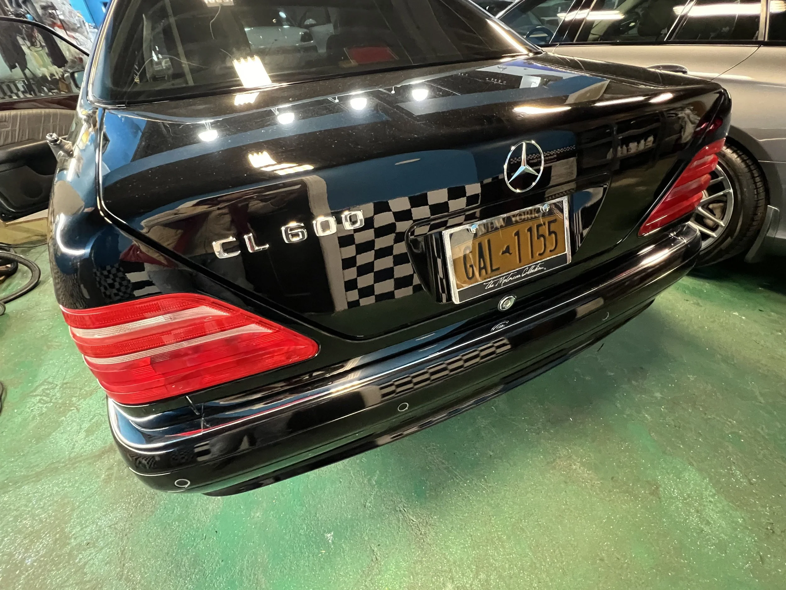 The back of a black Mercedes-Benz CL600 parked on a green floor, with a New York license plate reading GAL 1155. The car is in a showroom or garage with other vehicles visible nearby, and reflects overhead lighting.
