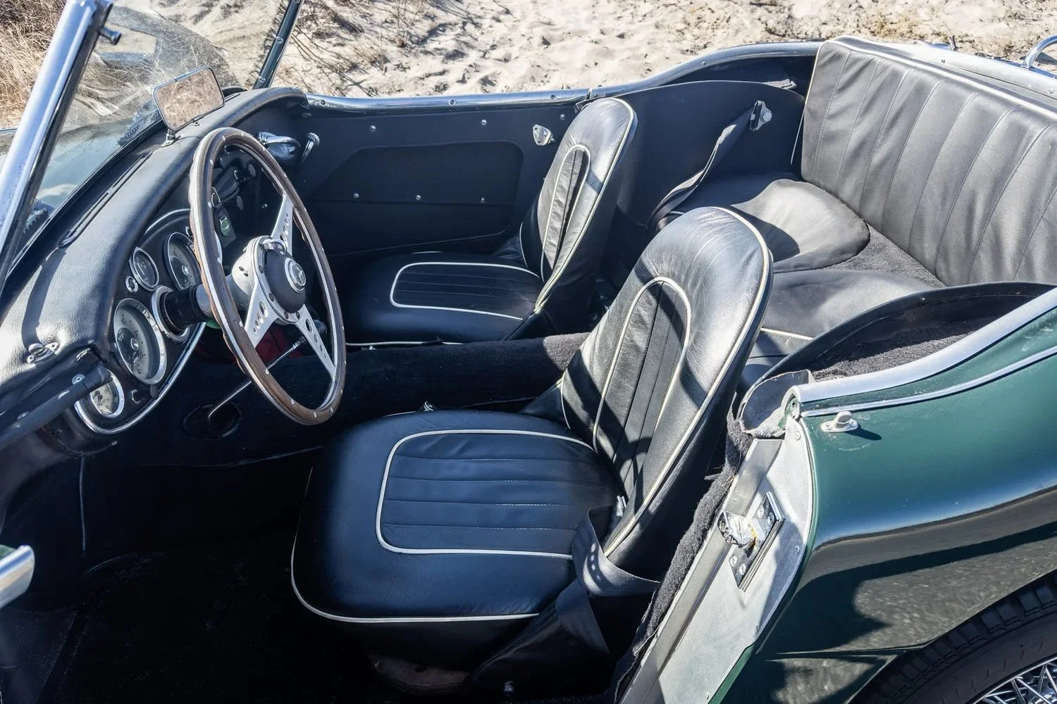 Interior of a classic convertible car with black seats, a wooden steering wheel, and vintage gauges, parked outdoors on sandy ground.
