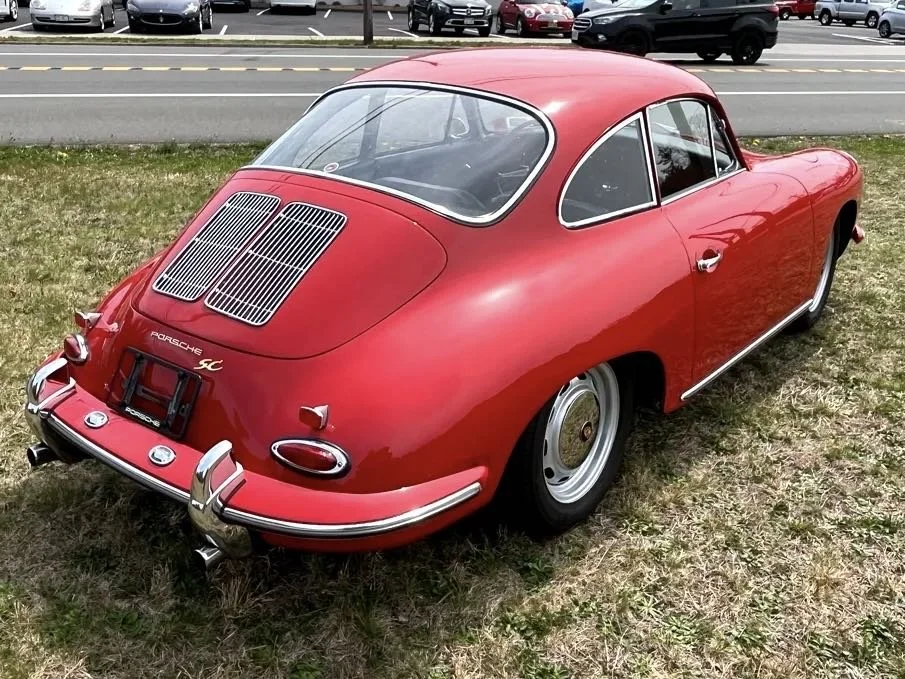 A vintage red Porsche 356 with a sleek, rounded body parked on a grassy area near a road, with cars and trees in the background.