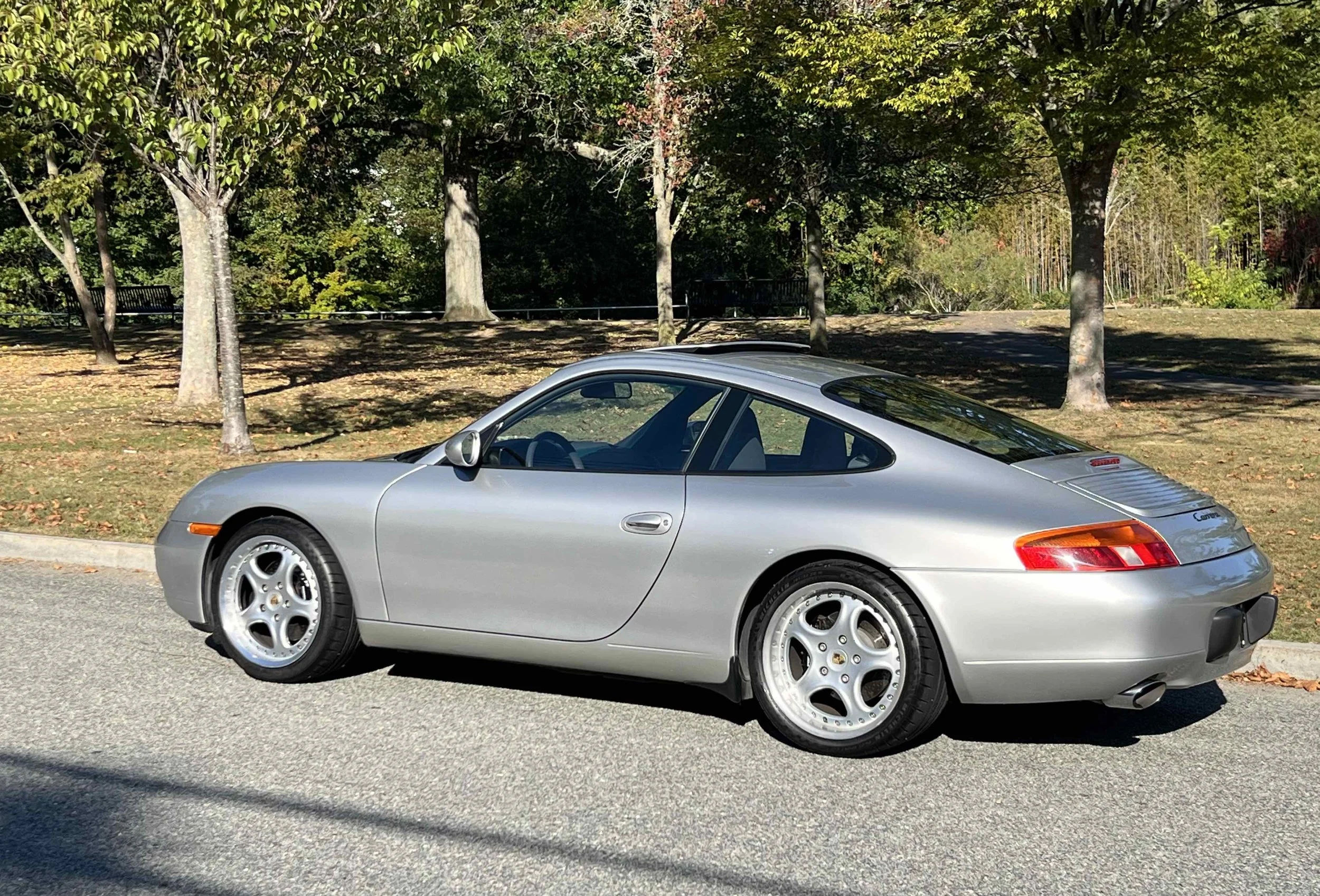 Silver Porsche 911 coupe parked on a street near a park with trees and fallen leaves, sunny day.