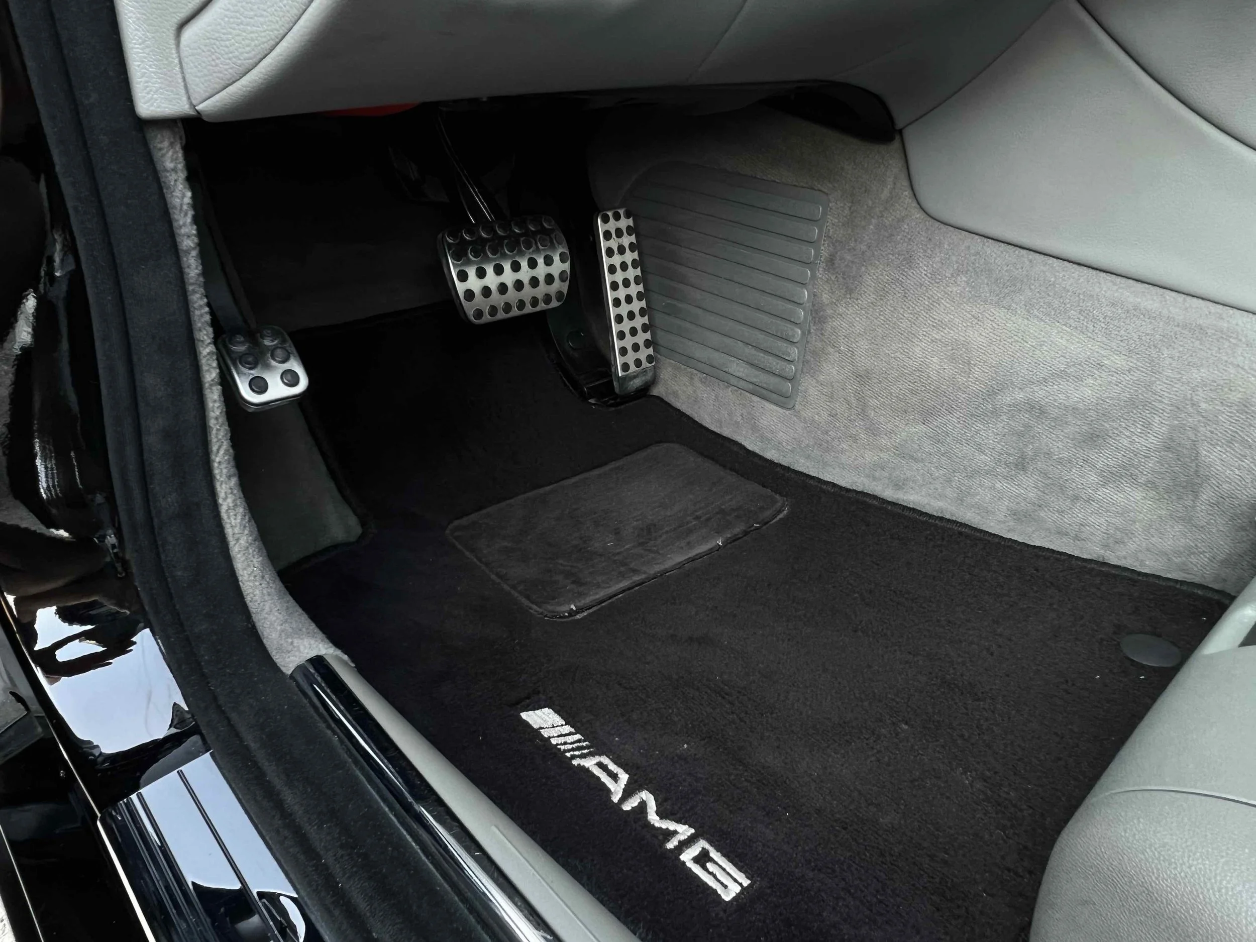 Interior car footwell showing AMG branded black carpet, three metallic pedal covers with circular holes, and a light-colored side panel with a rubber footrest.