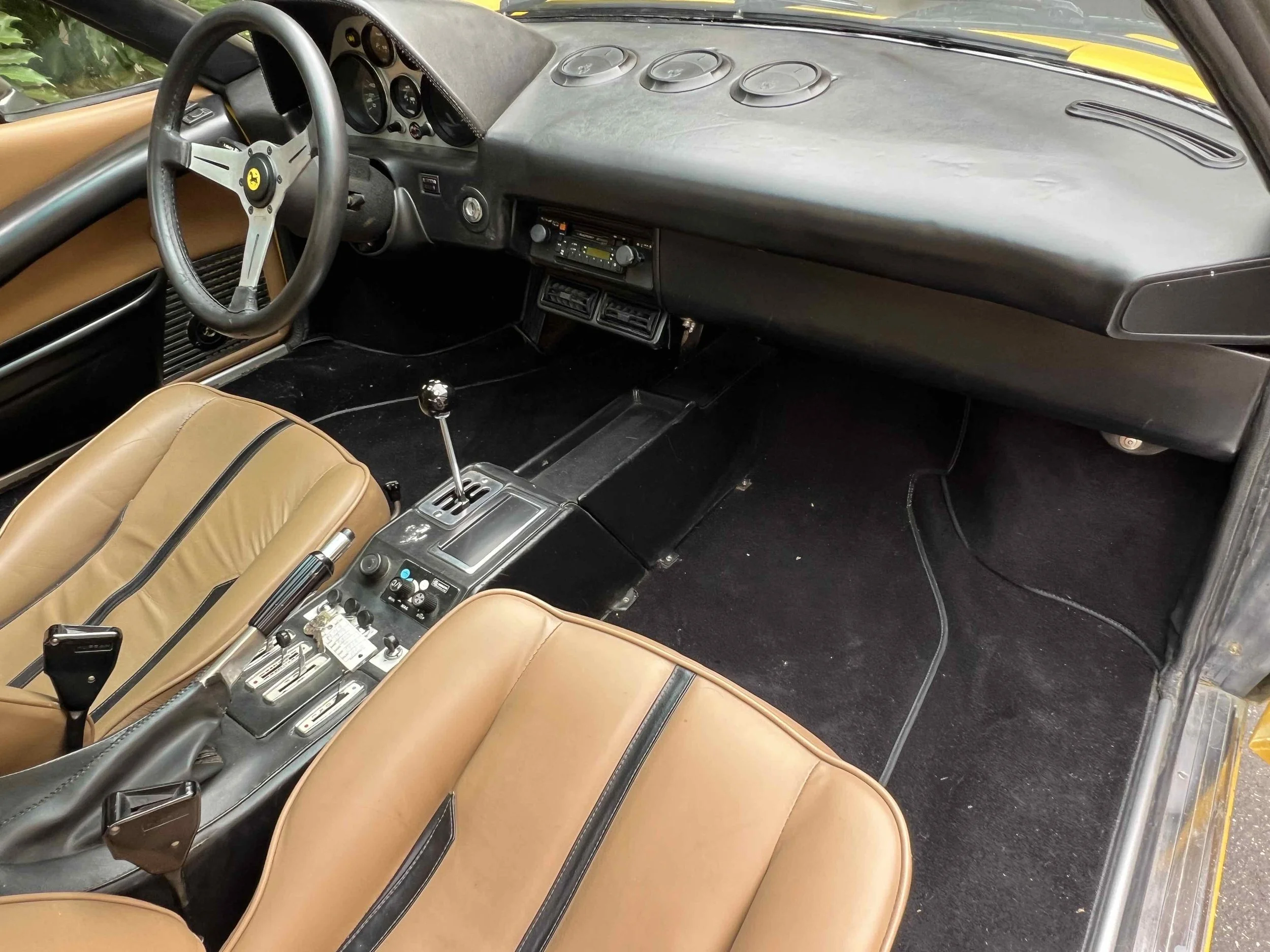 The interior of a classic car with tan leather seats, a black dashboard, and a manual gear shift. The steering wheel features a Ferrari logo.