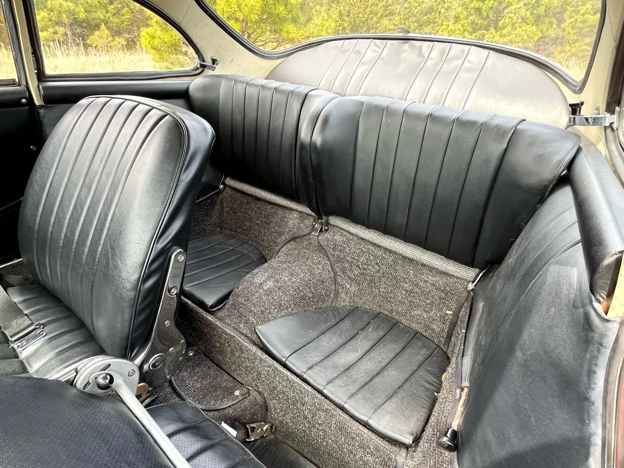 The interior of a vintage car featuring black leather seats, with two front seats and a rear bench seat, black carpeting, and large side windows, with a background of greenery seen outside.