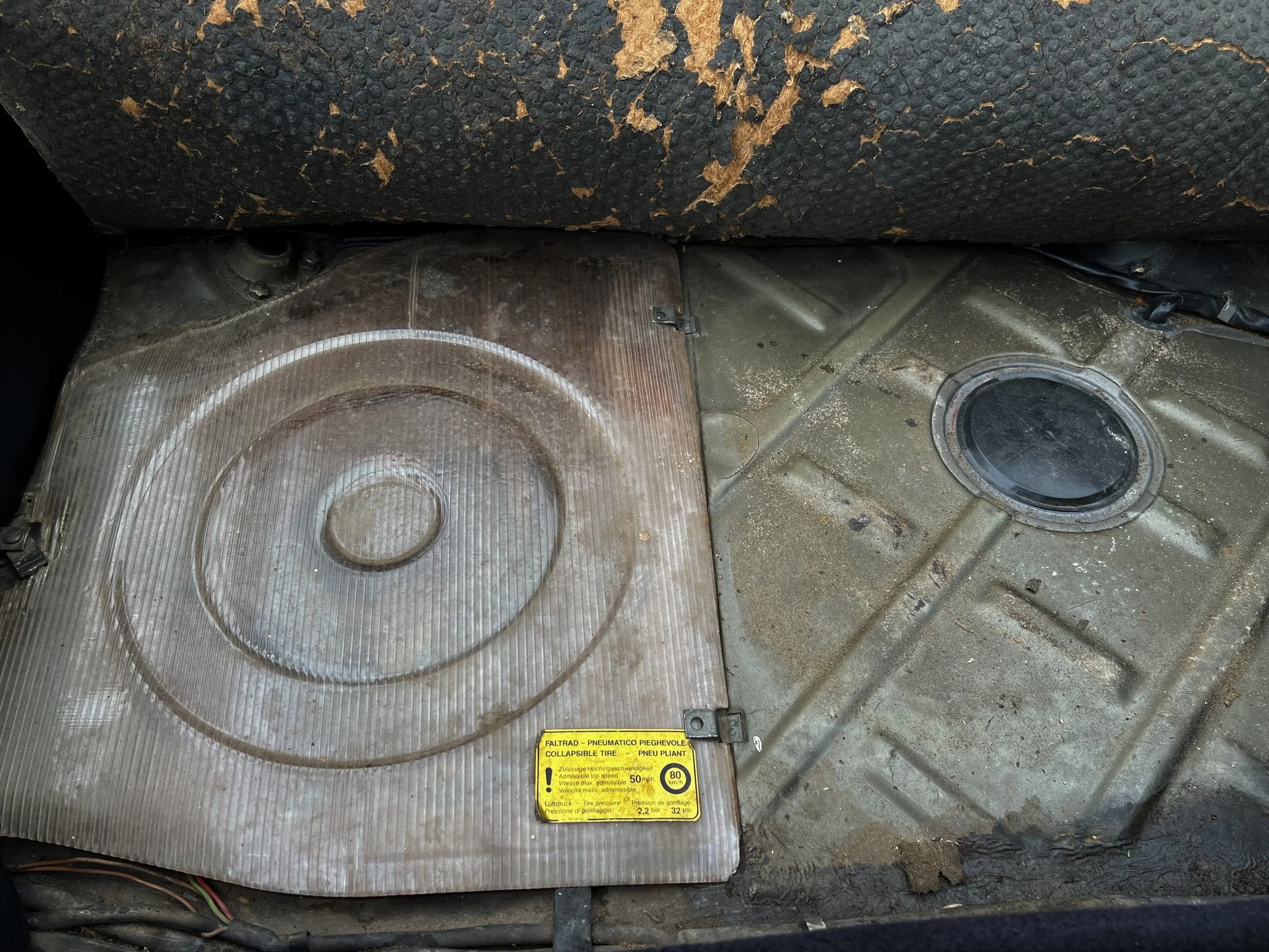 Dirty, worn metal surface inside a vehicle, with a round black rubber plug or cap on the right side and a yellow warning label on the left side.