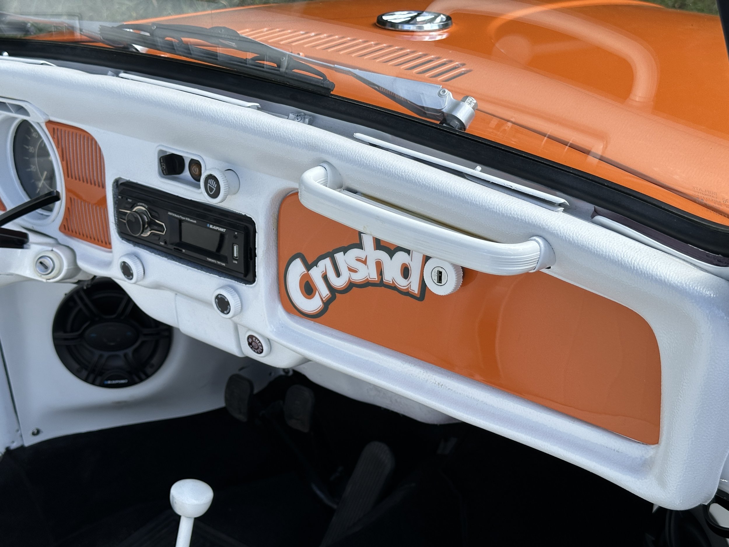 Interior of a vintage Volkswagen van, featuring a white dashboard with orange accents and a sticker that says 'crushd' in orange letters.