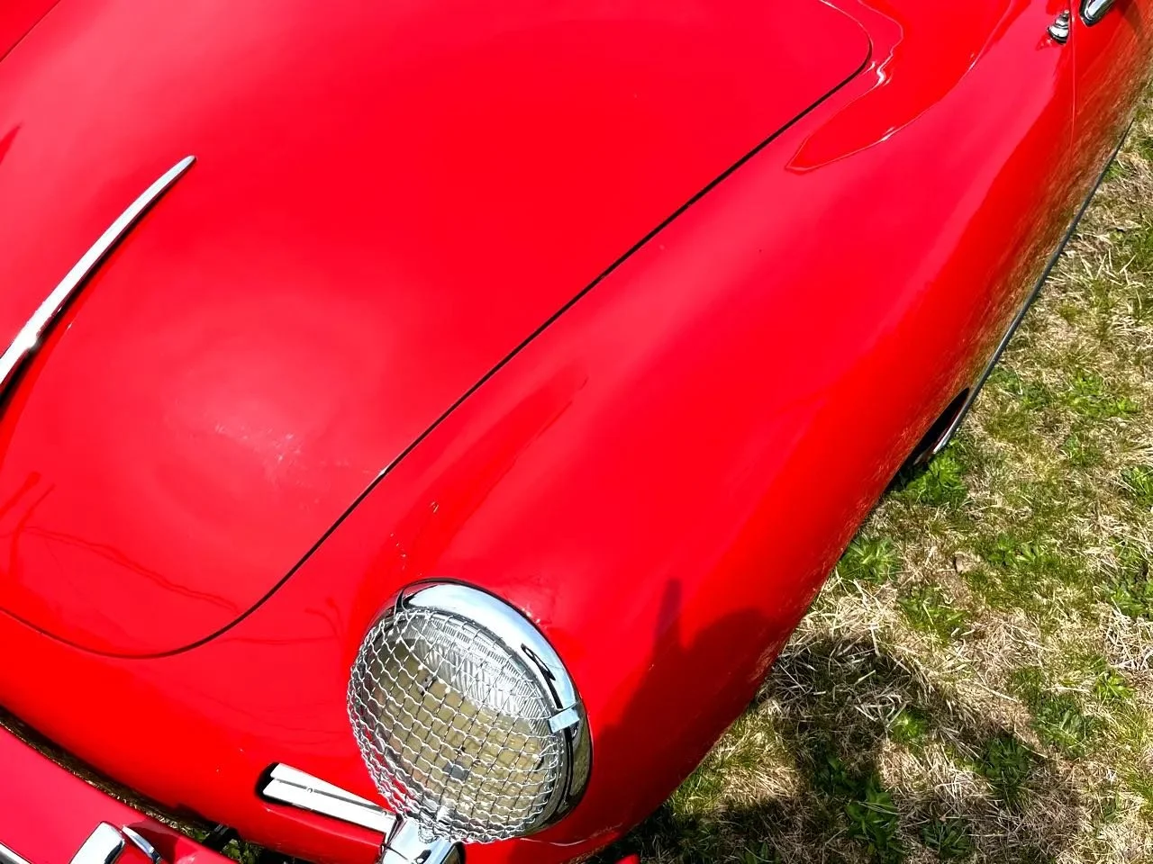 Close-up of a vintage red car's front left side, featuring a chrome headlight with a grille cover, a curved body panel, and part of the hood, with the car parked on grass and dirt.