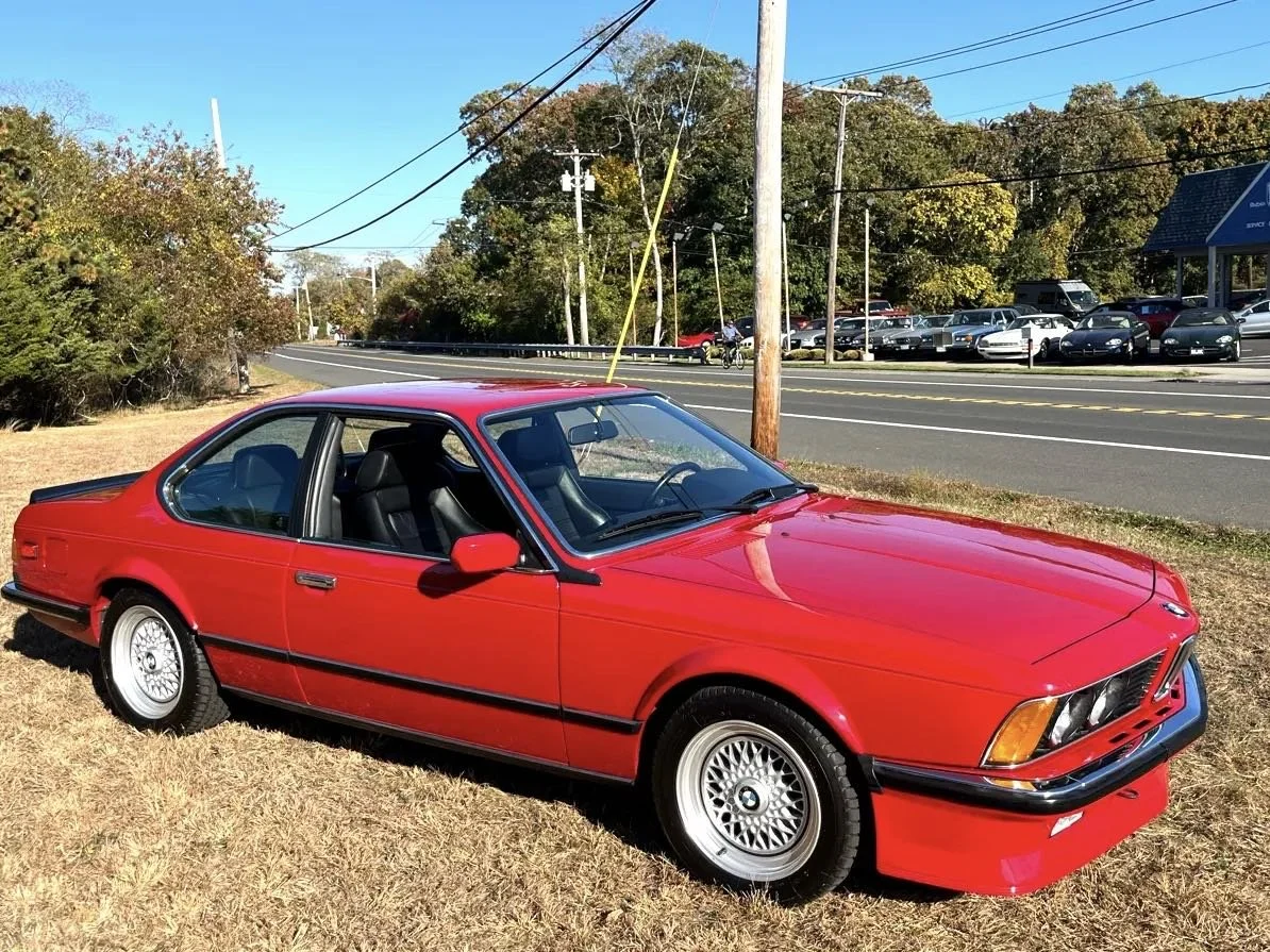 Red vintage BMW coupe parked on a grassy area beside a road with trees and a parking lot in the background.