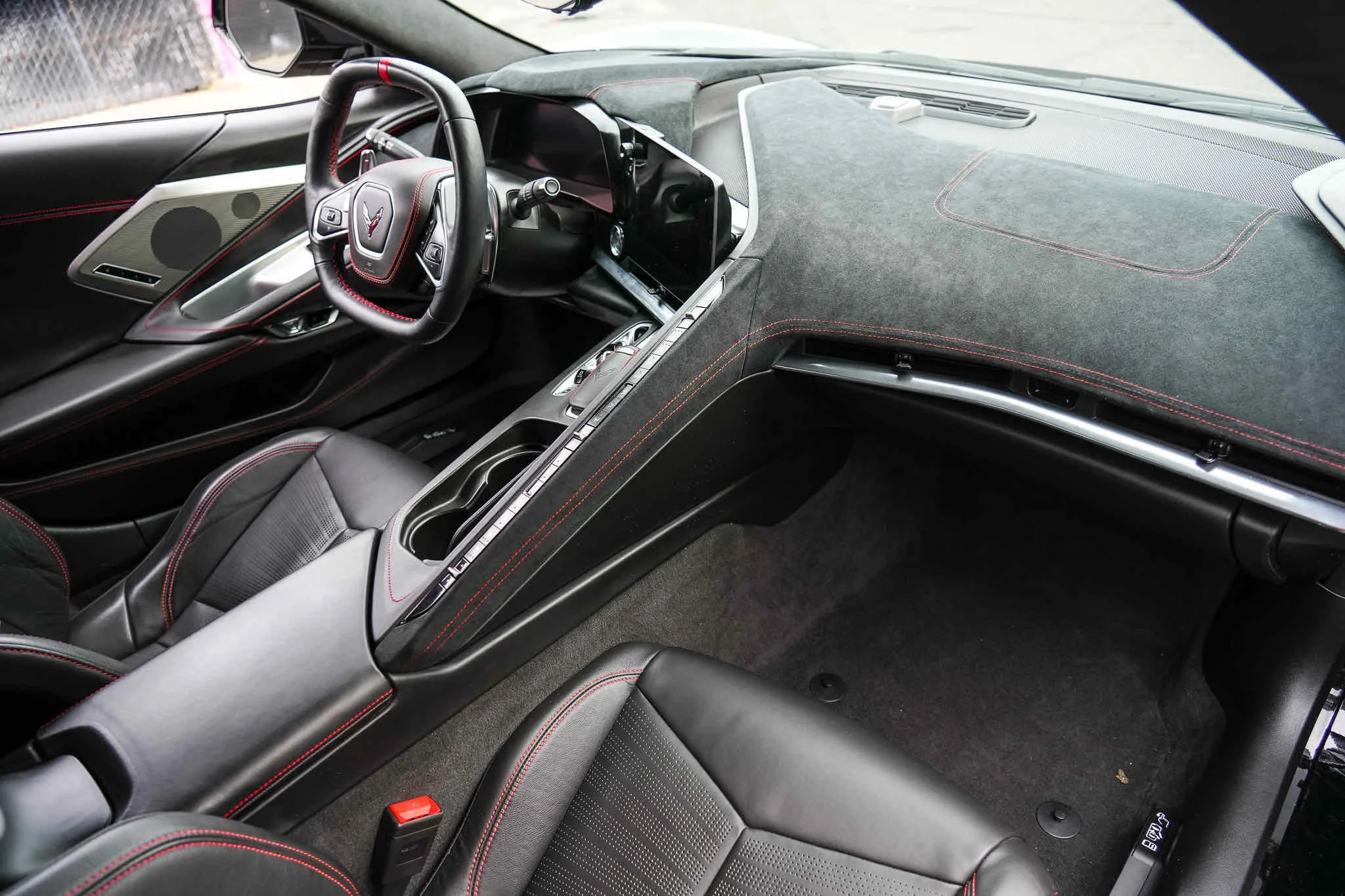 Interior of a modern sports car, showing the steering wheel, dashboard, center console with cup holders, and black leather seats with red stitching.