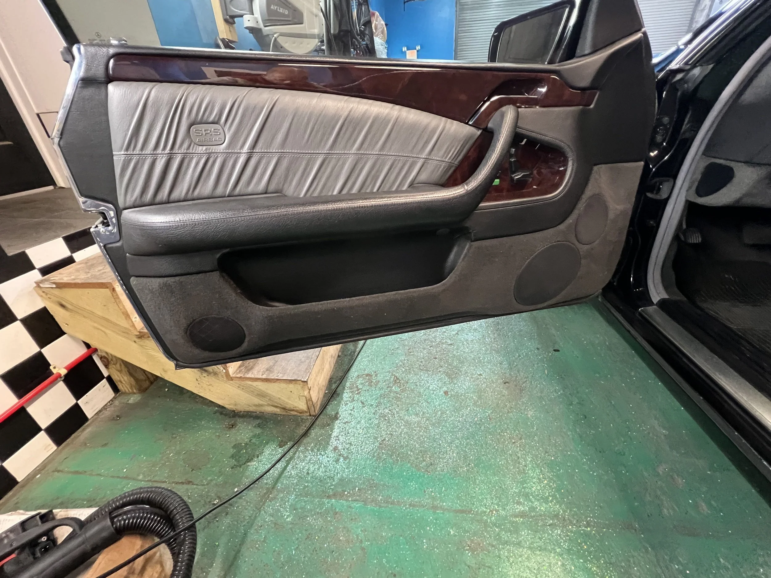 Car door interior with leather and wood trim, speaker, window controls, and an armrest, in a garage setting.