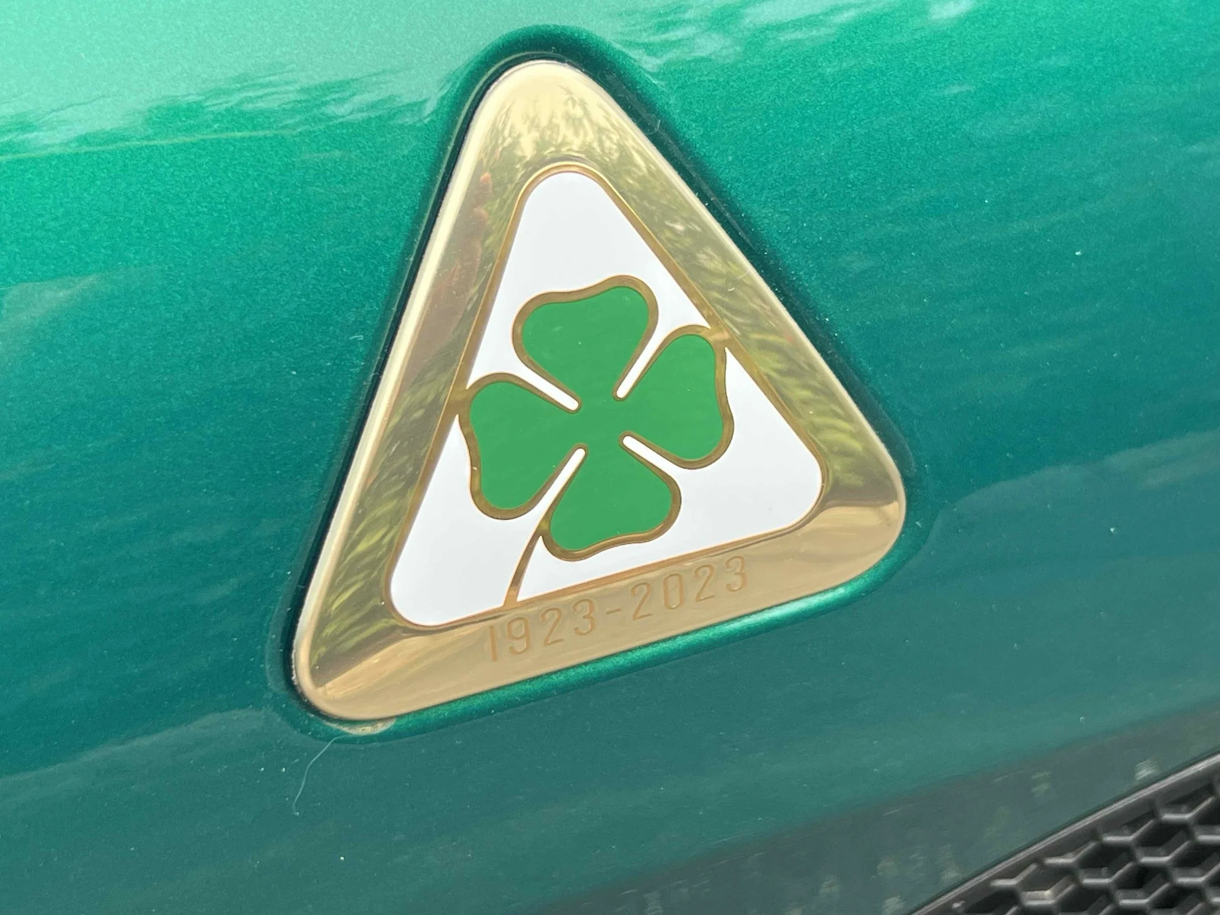 A triangular emblem with a green four-leaf clover symbol, gold border, on a green surface, with the years 1923-2023 engraved at the bottom.