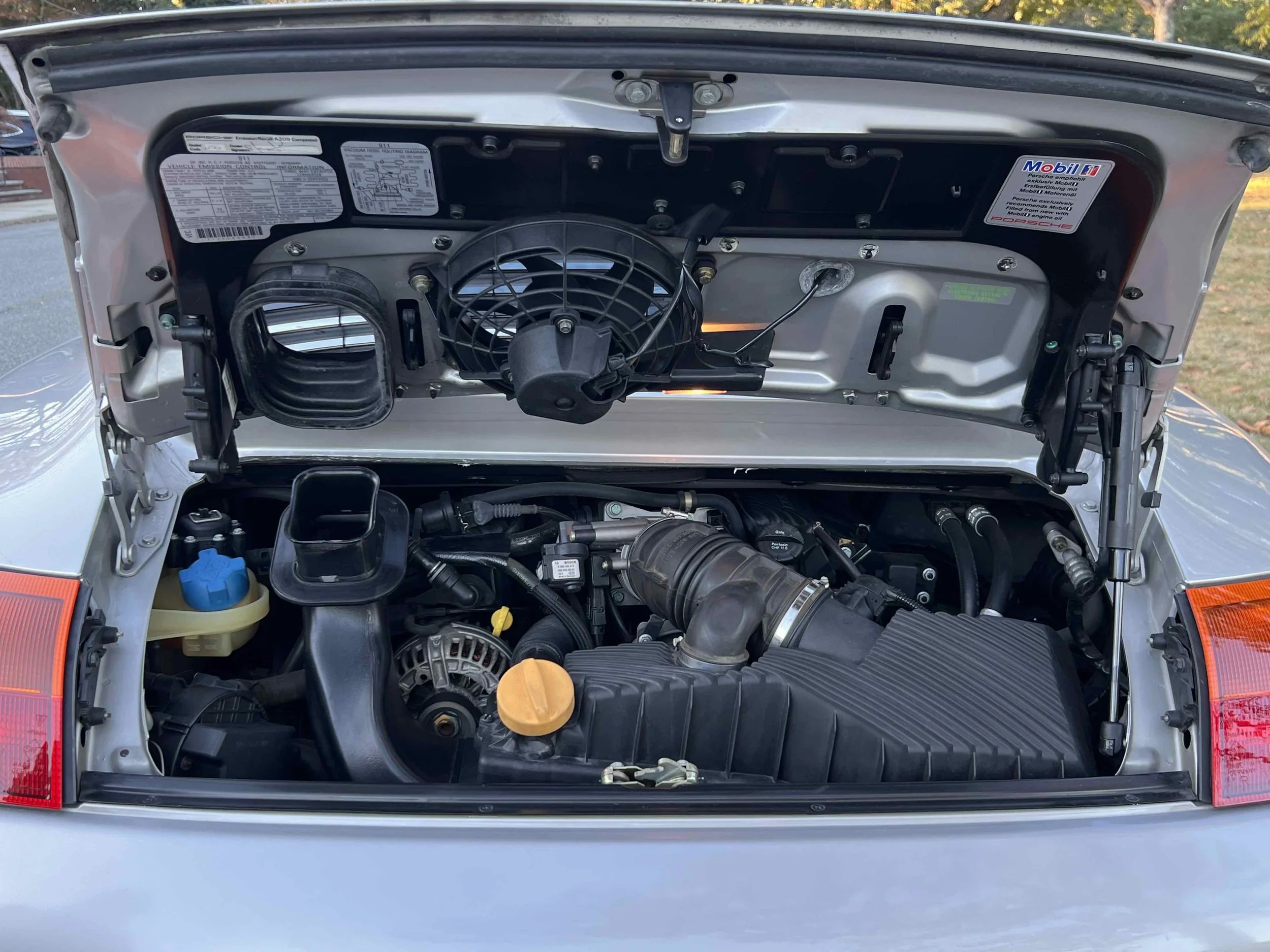 View of the engine bay of a silver sports car with the hood open, showing various engine components, hoses, and labels.