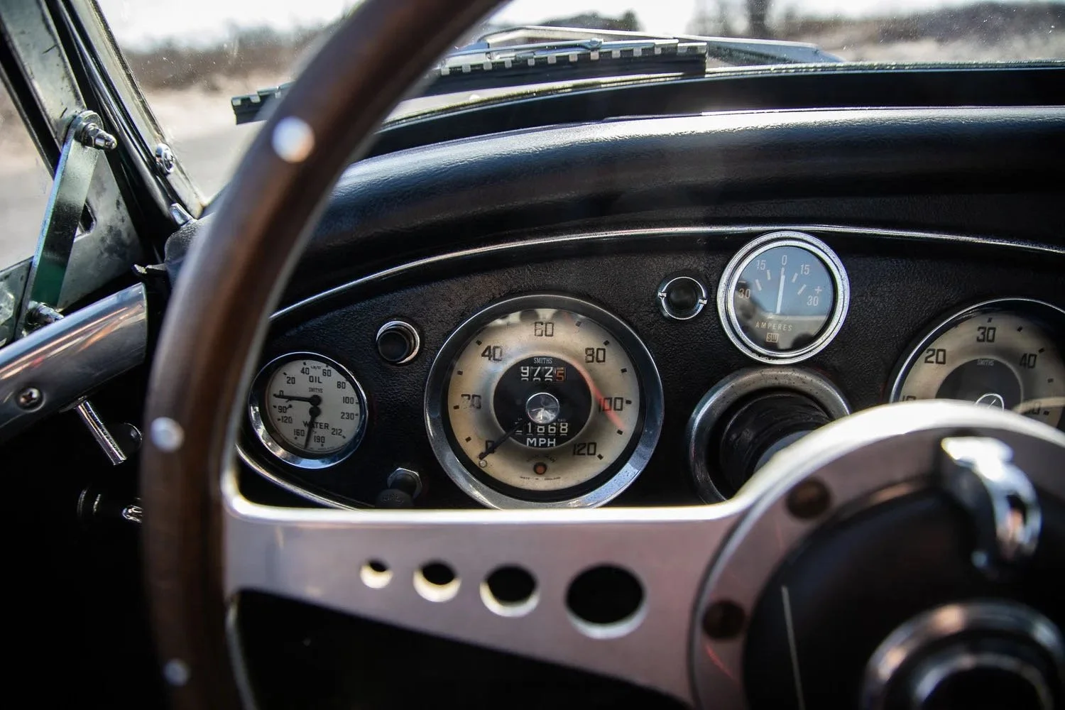 Dashboard of a vintage car showing speedometer, tachometer, oil and water temperature gauges, and ammeter, with windshield and blurred landscape in the background.