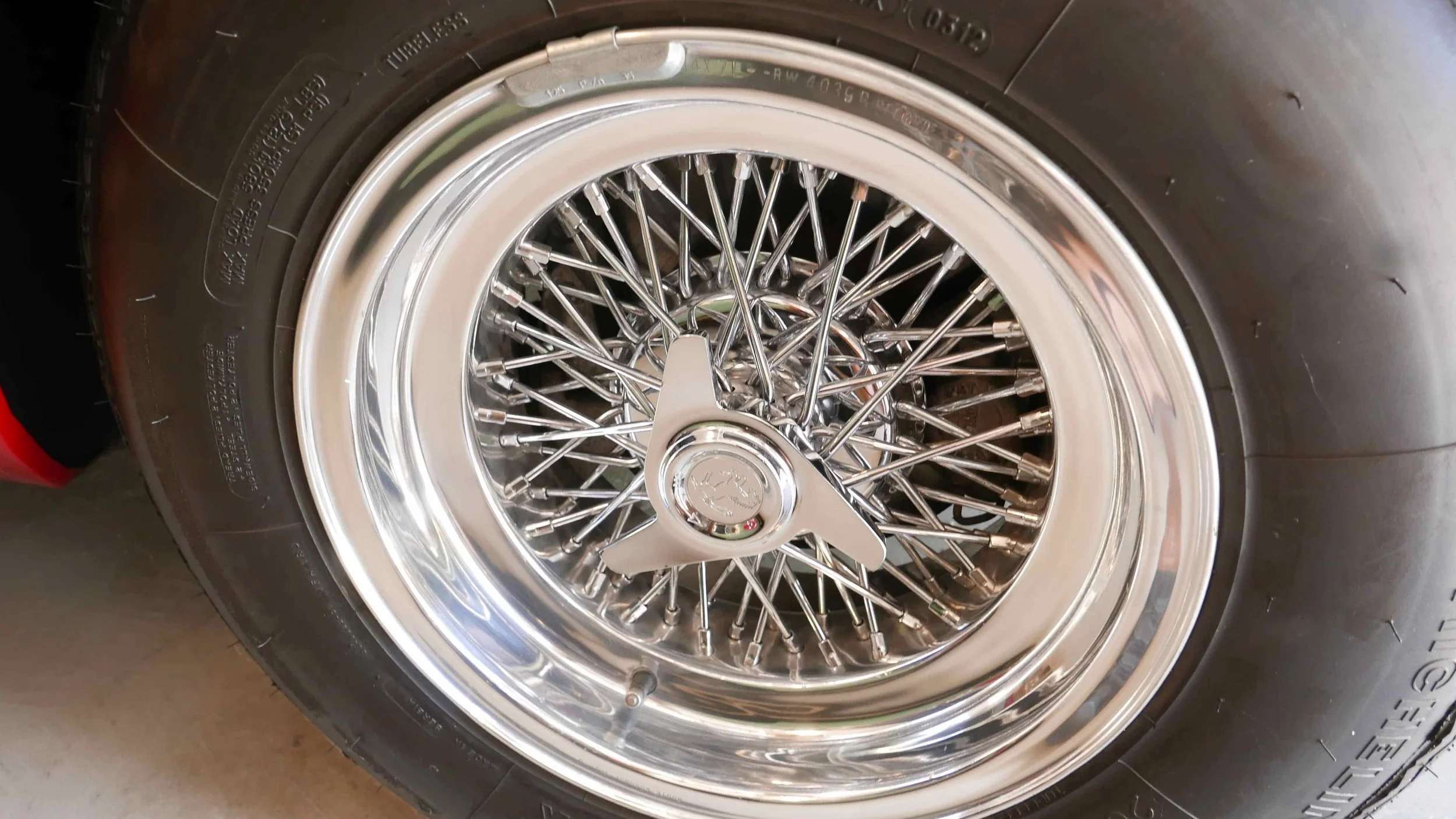 Close-up of a vintage wire-spoke car wheel with a chrome finish. The wheel is fitted with a black tire and features a hubcap with a small logo or emblem in the center.