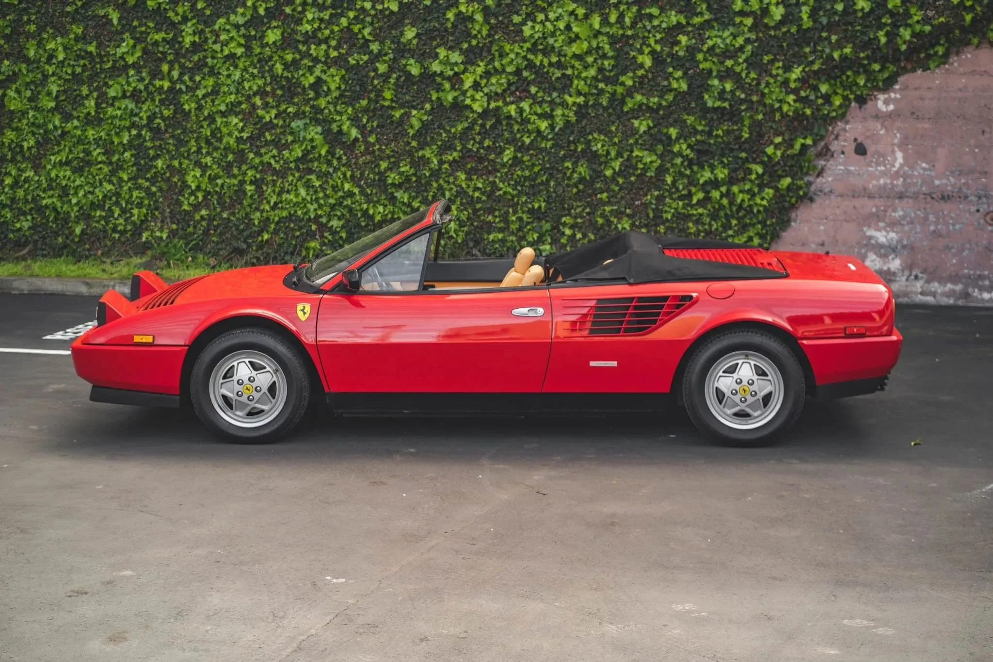 Red Ferrari convertible parked on the street with a green hedge and brick wall background.