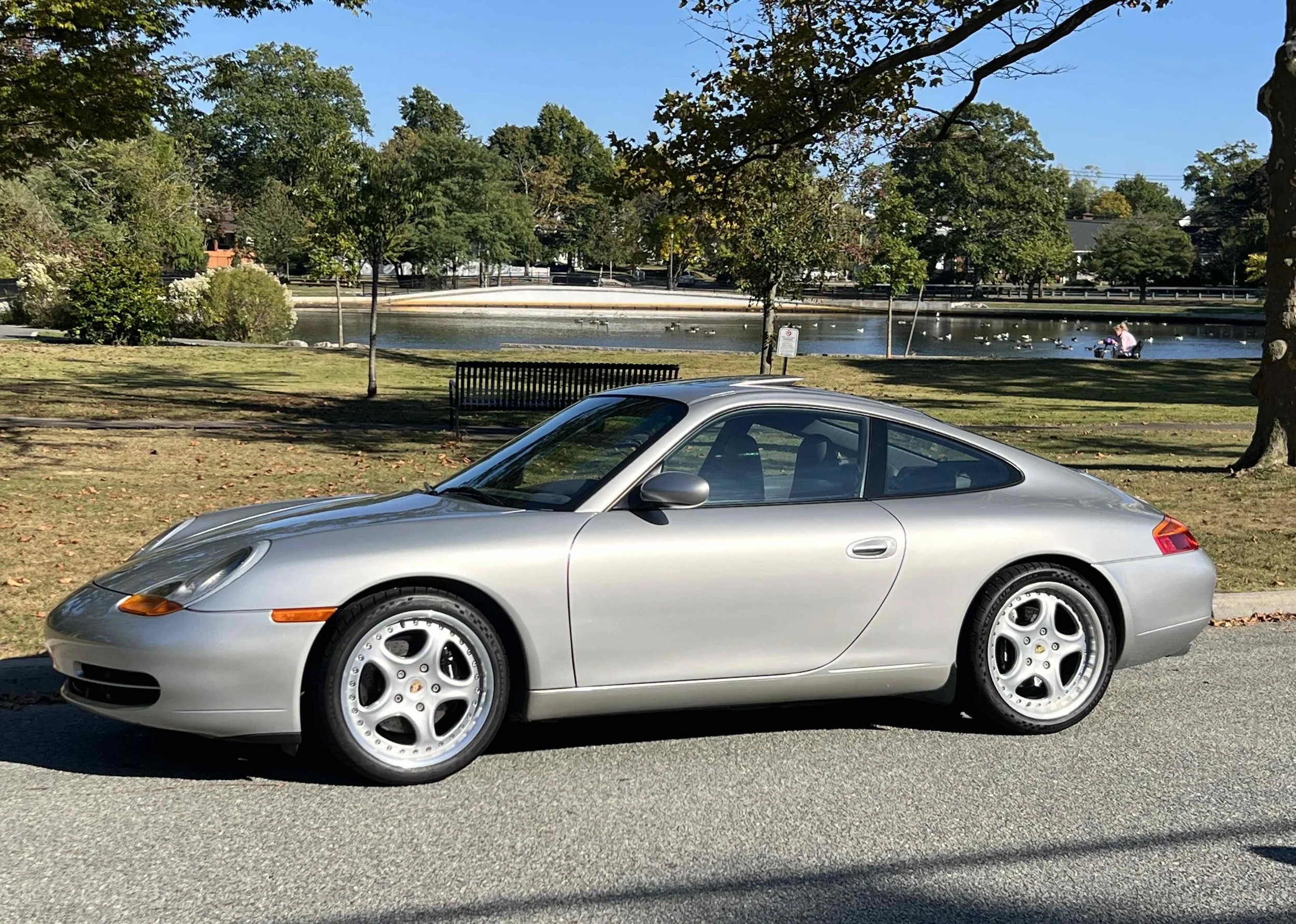 Silver Porsche 911 parked on a street near a park with trees and a pond, people in the background
