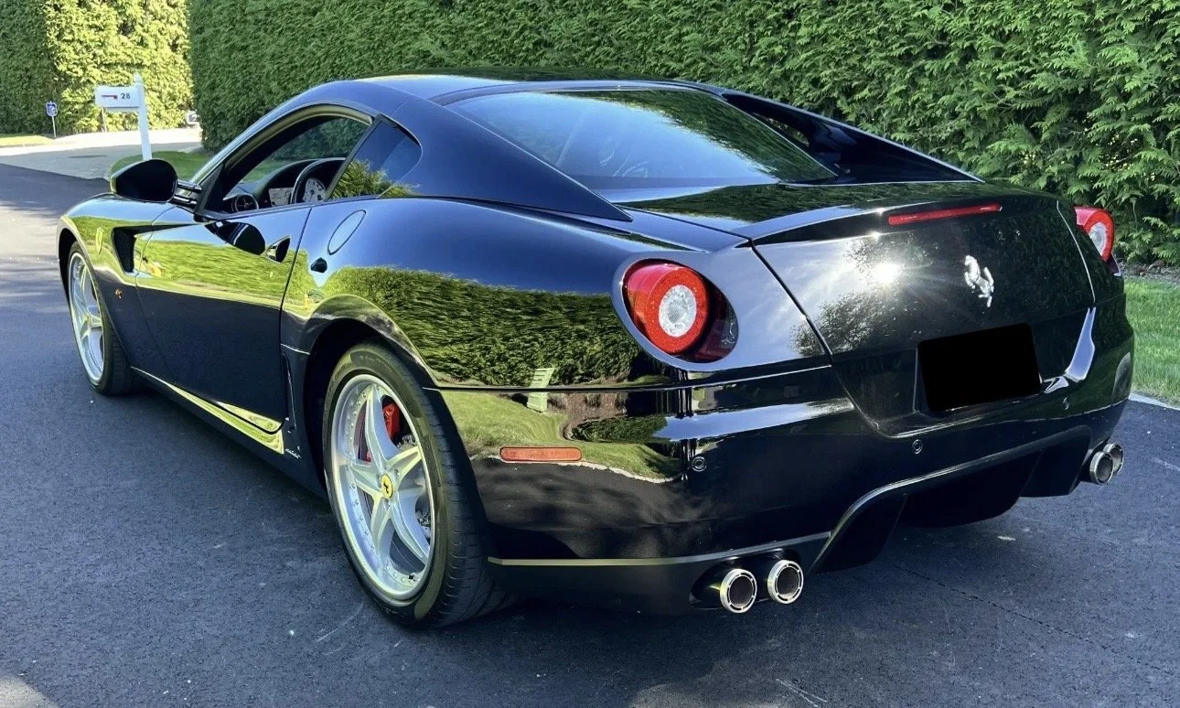 Black Ferrari sports car parked on a paved driveway with green trees in the background.