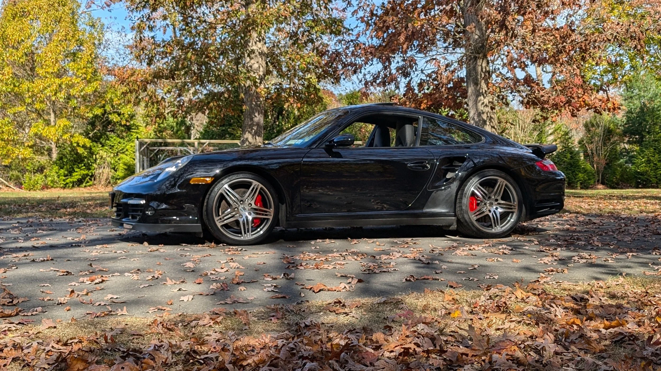 Black sports car parked on a leaf-covered road with autumn trees in the background.