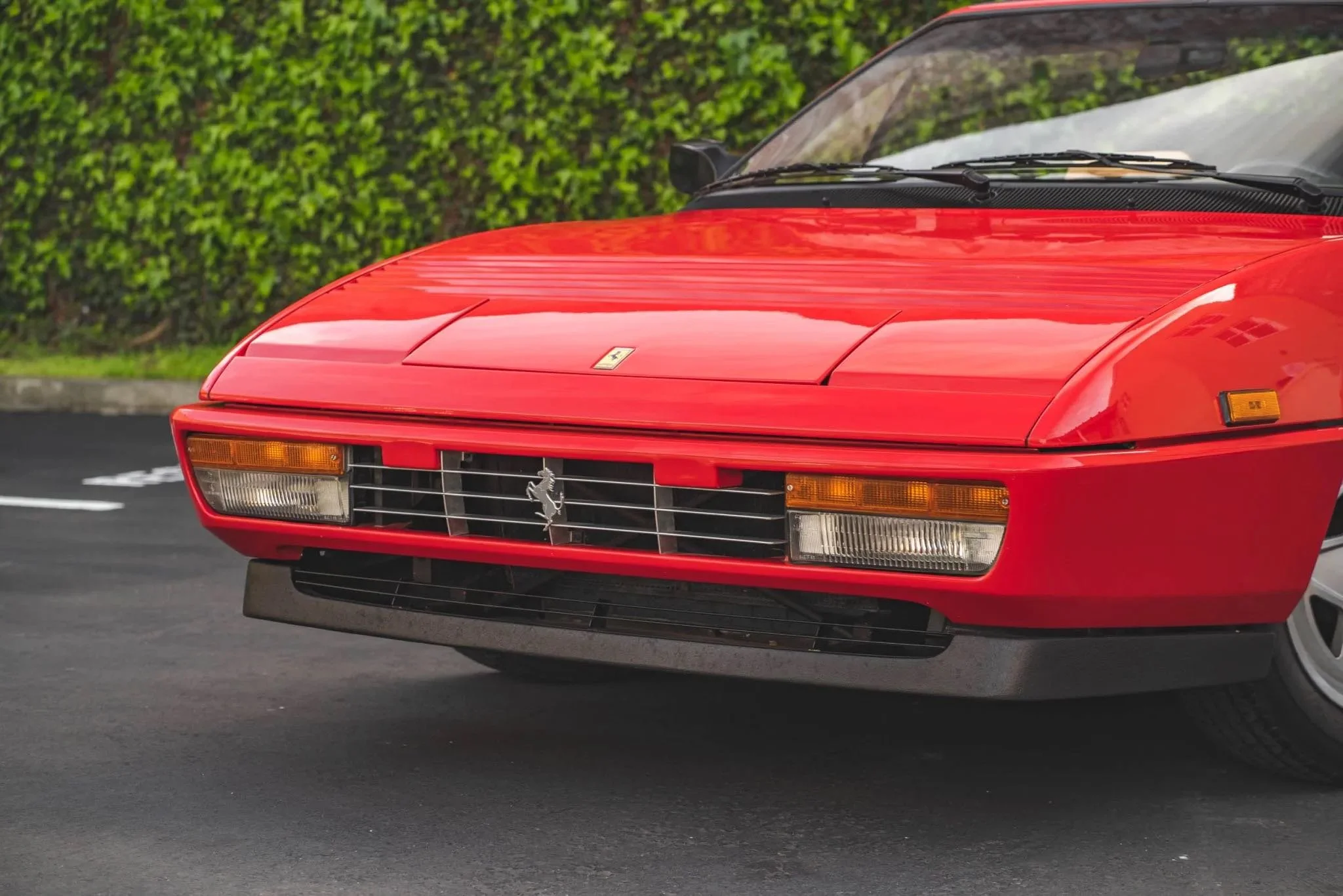 Close-up of the front end of a red classic Ferrari sports car, showing the grille with a Ferrari emblem, rectangular headlights, and a sleek hood.