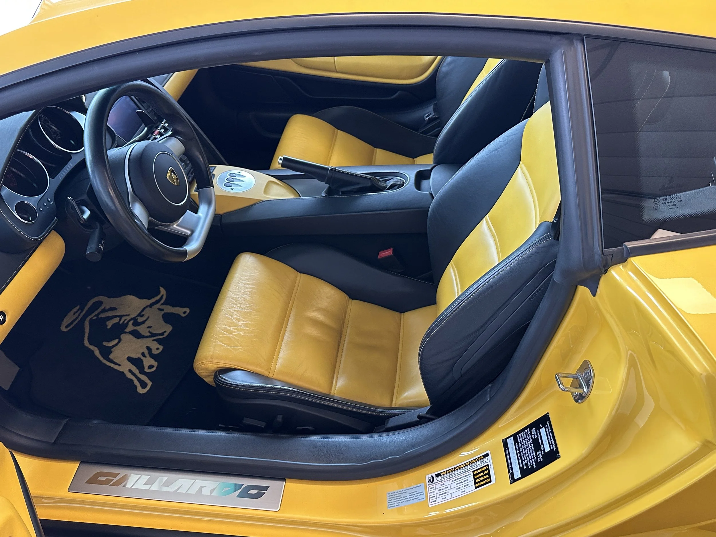 Interior of a yellow Lamborghini Gallardo with black and yellow leather seats, black steering wheel, and black floor mat with a lion head logo.