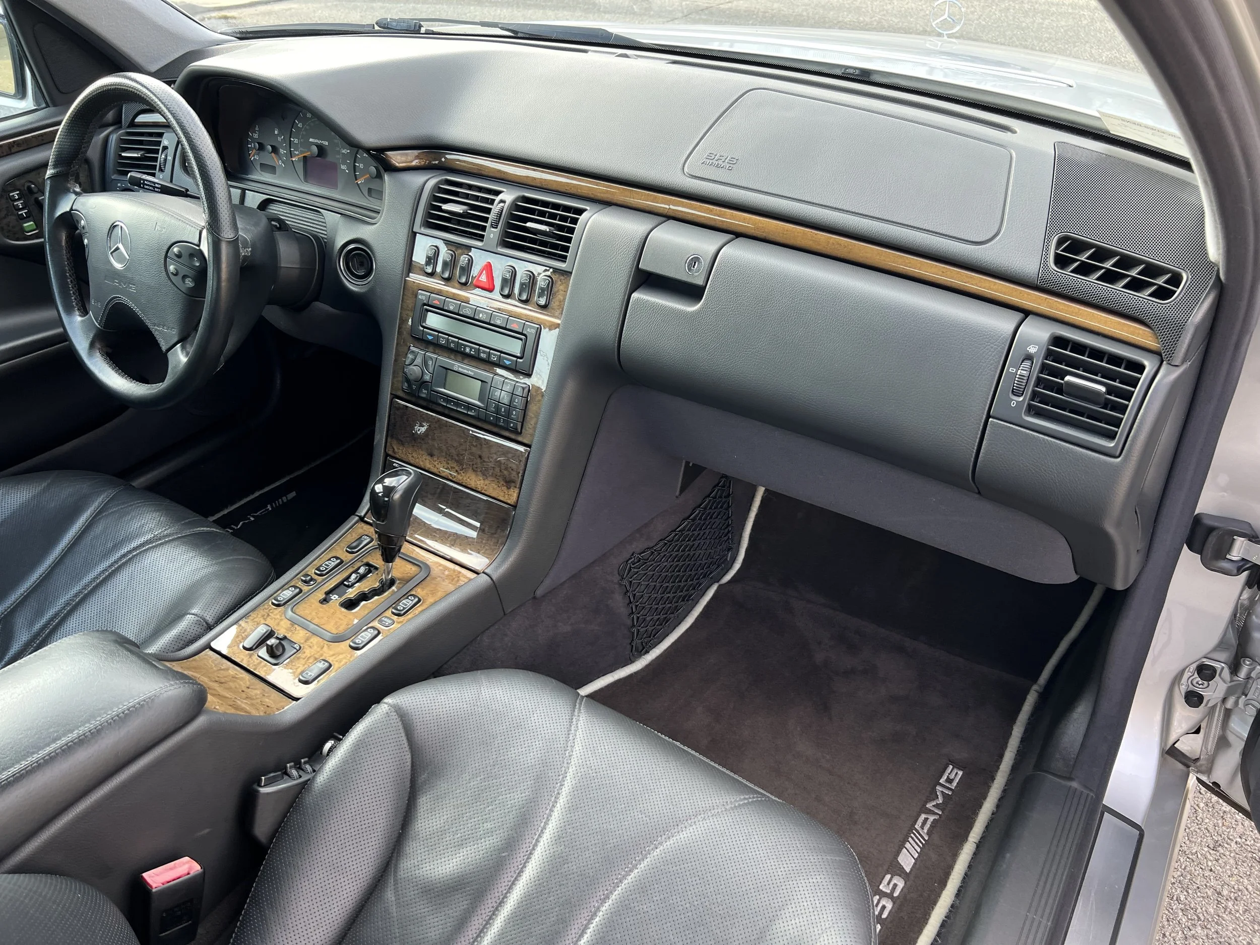 The interior of a Mercedes-Benz vehicle, showing the dashboard, steering wheel, center console with wood trim, gear shift, and black leather seats.