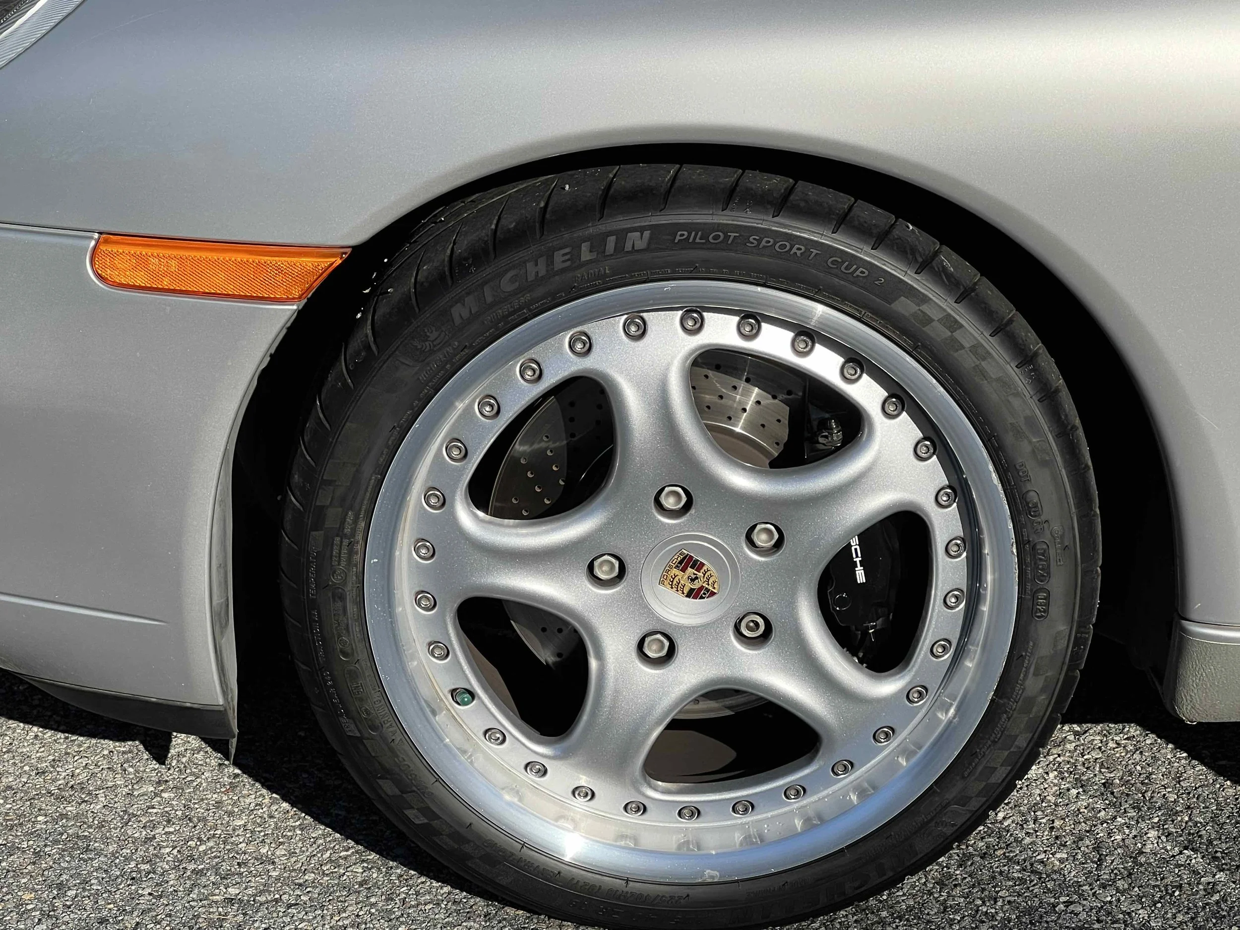 Close-up of a silver Porsche car wheel with a Michelin Pilot Sport Cup 2 tire, a multi-spoke alloy rim, and a Porsche badge center cap.
