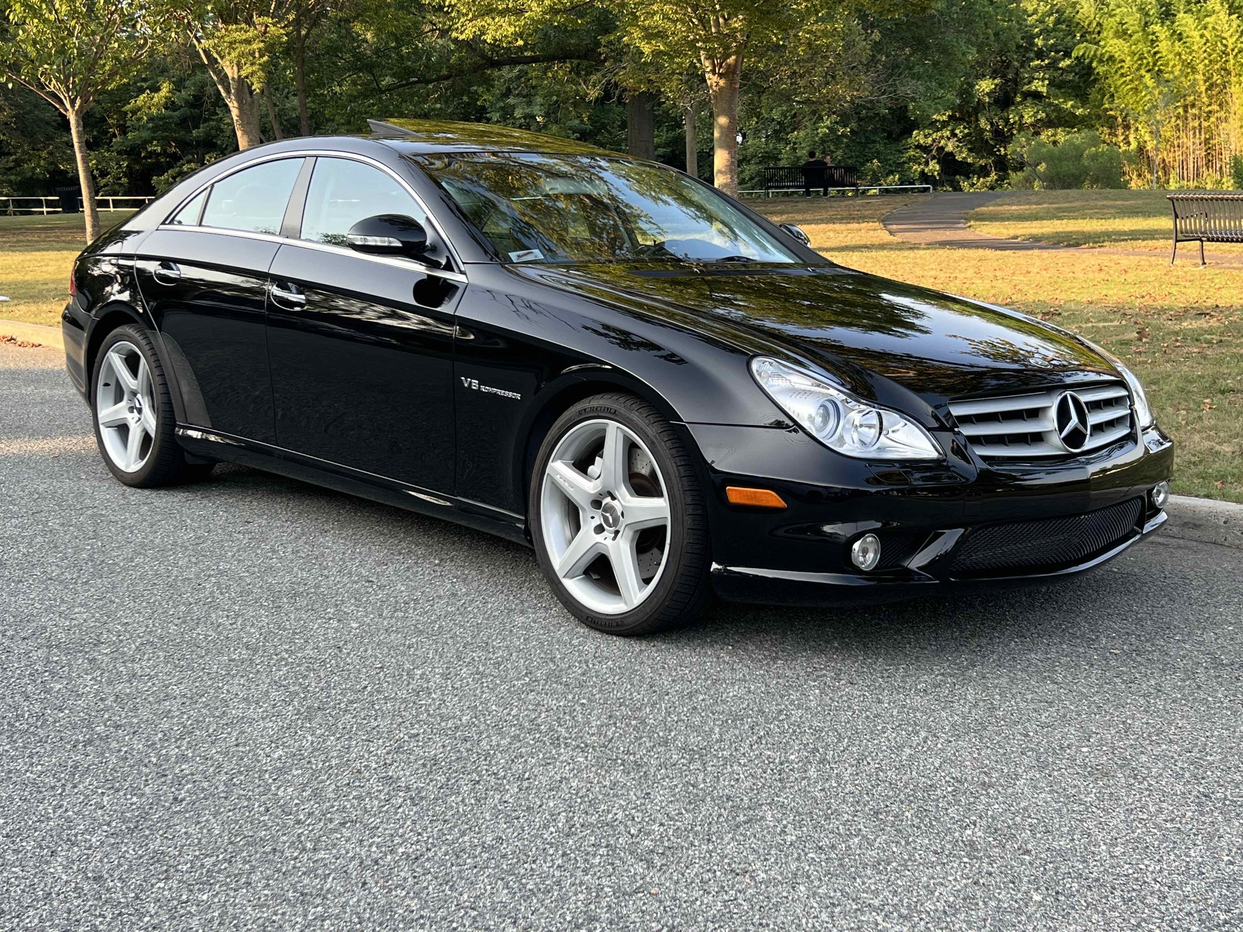 Black Mercedes-Benz CLS55 AMG  parked on a street in a park with trees and benches in the background.