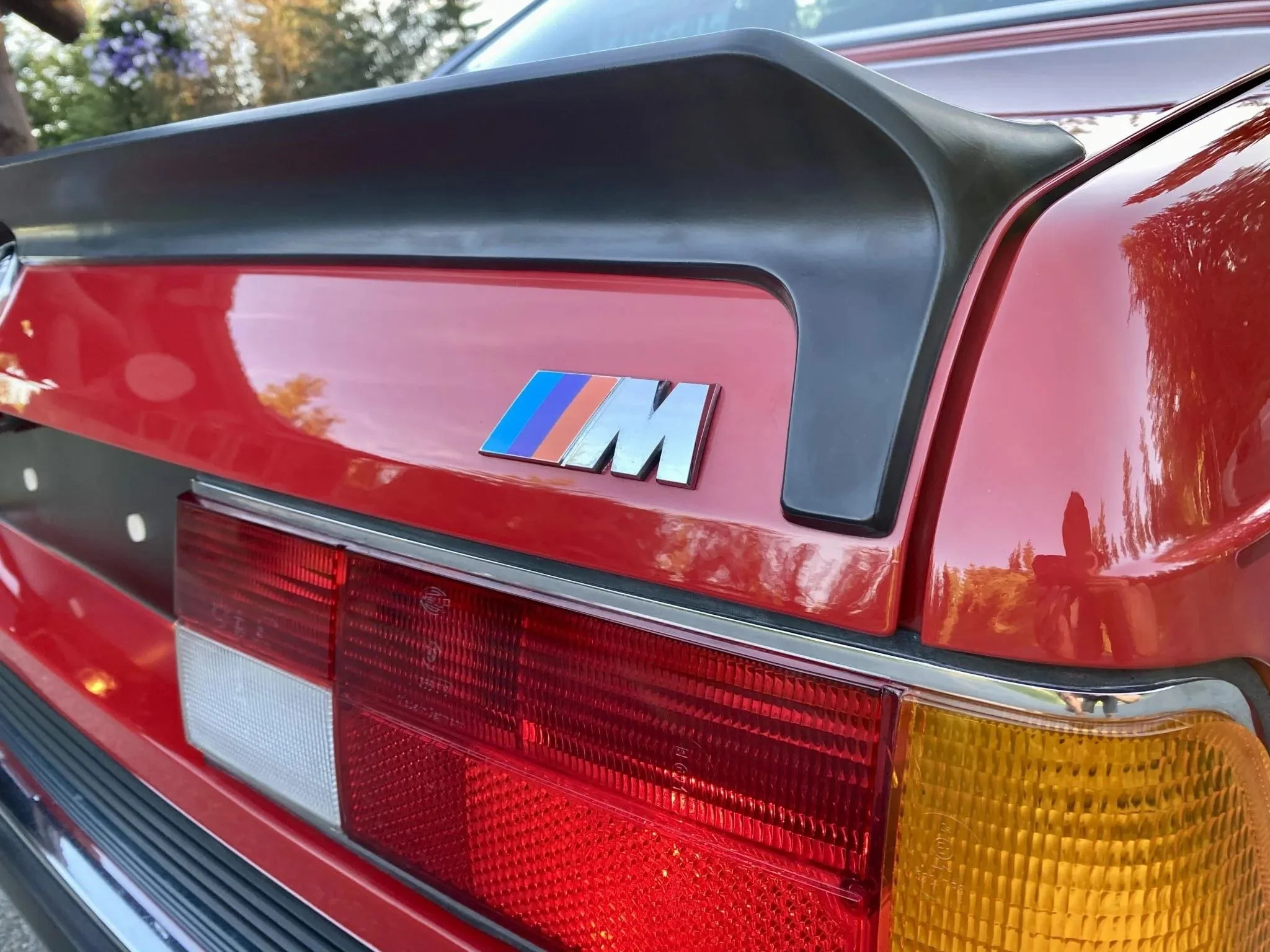 Close-up of the rear end of a vintage red BMW M car, showing the taillight, black spoiler, and M badge.
