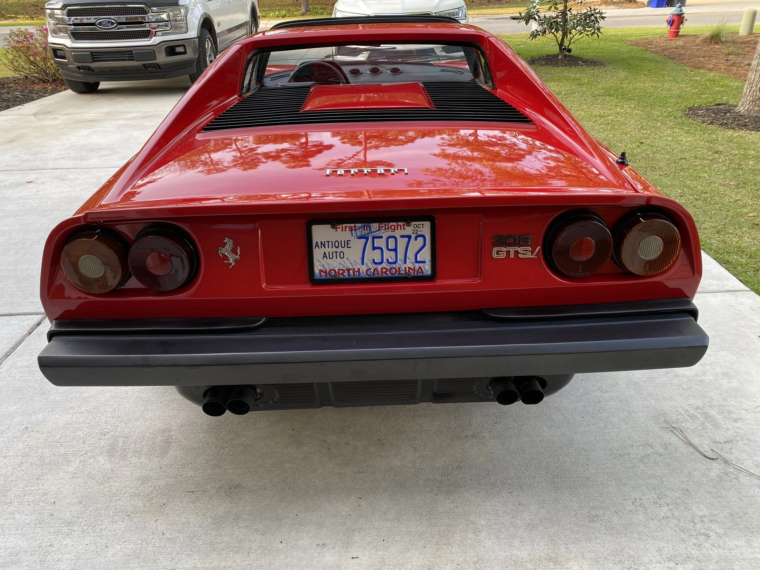 The rear view of a classic red Ferrari 308 GTS/ in a driveway with a North Carolina license plate, flanked by a silver pickup truck on the left and a grassy lawn with trees and a fire hydrant on the right.