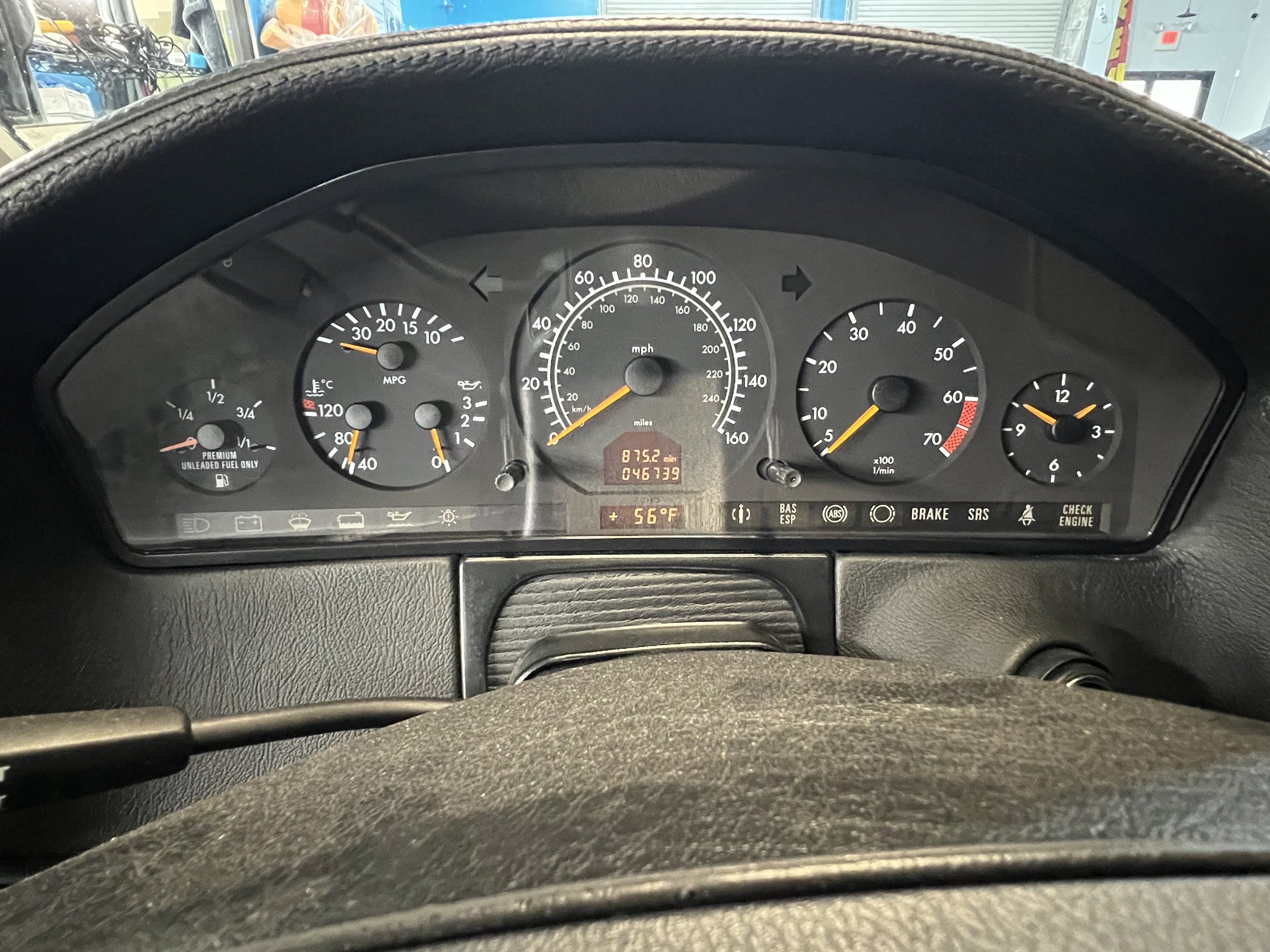 Dashboard of a car showing speedometer, fuel gauge, temperature gauge, tachometer, and various warning lights, with a background of a garage.
