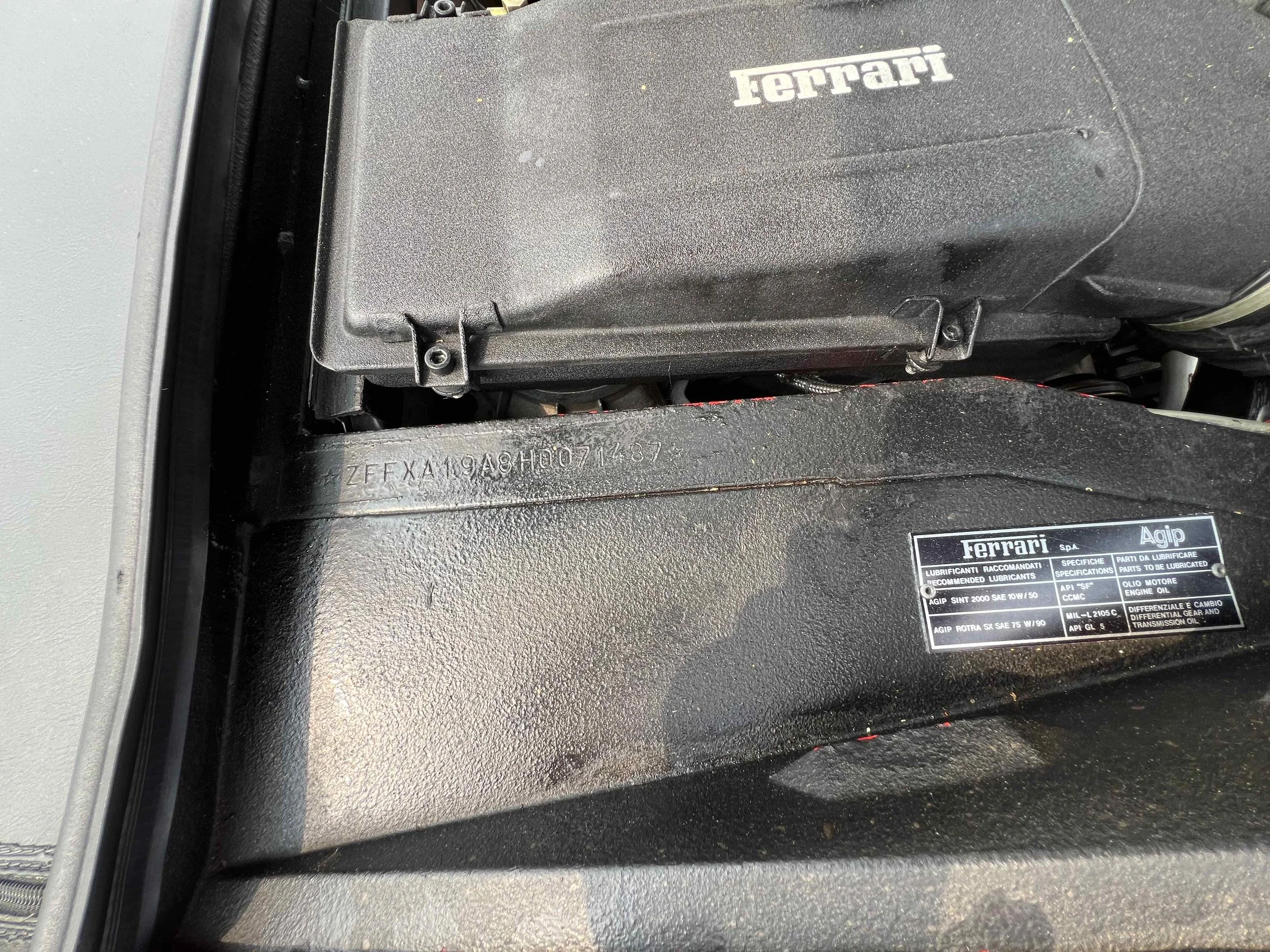 Close-up of a Ferrari engine compartment showing a black cover with the Ferrari logo and a label with technical specifications inside a vehicle.