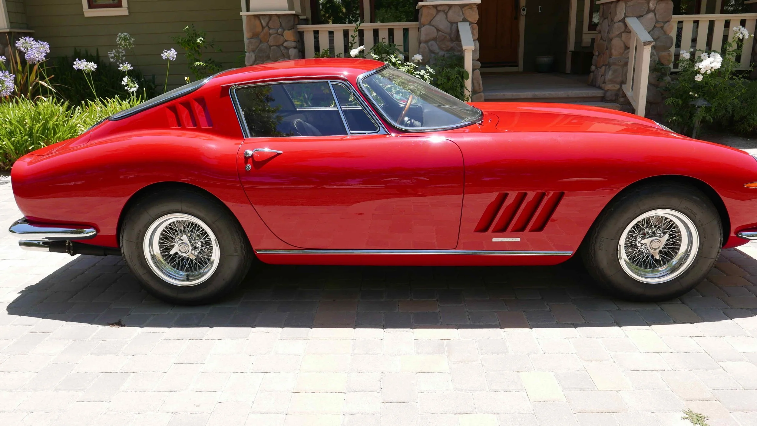 Red vintage sports car parked on a driveway, with a house and garden in the background.