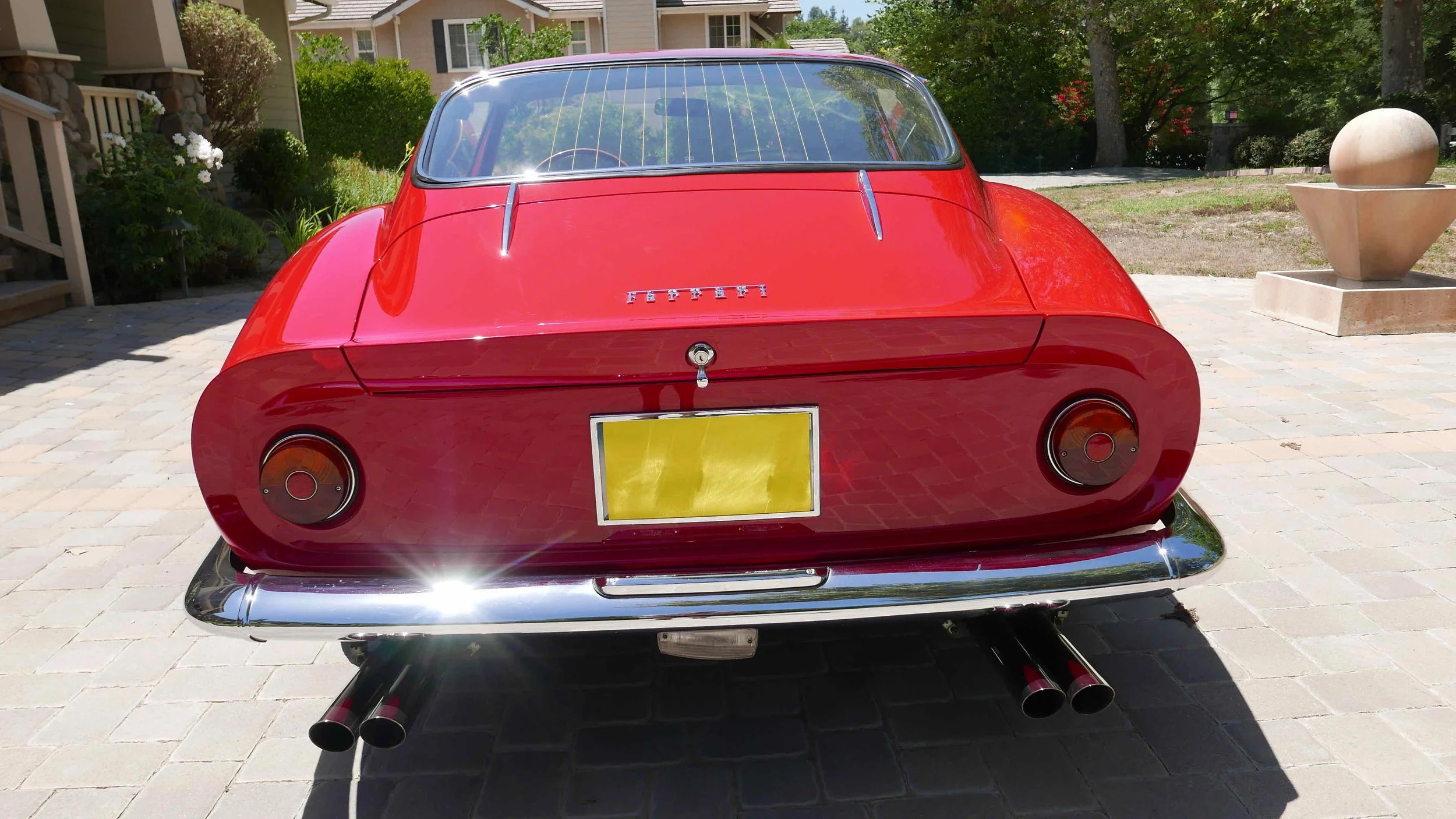 Red vintage Ferrari sports car parked on a stone driveway with house and garden in background.