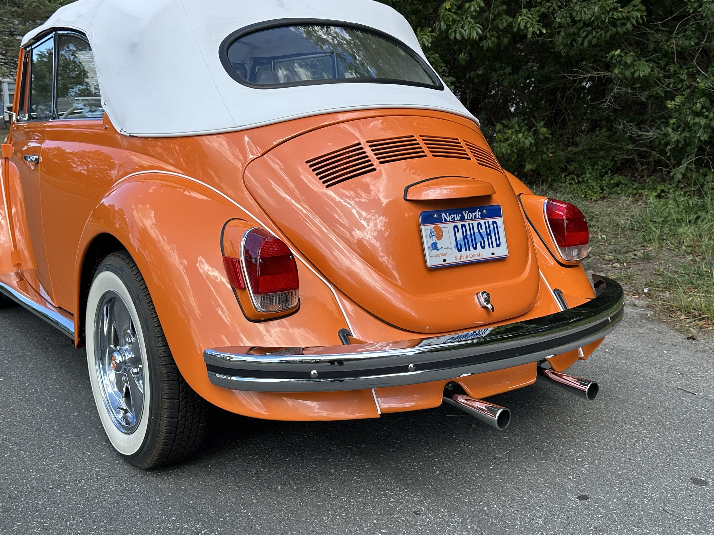 Rear view of a vintage orange Volkswagen Beetle convertible with a white soft top, dual exhaust pipes, and a New York license plate reading 'CRUSHD.'