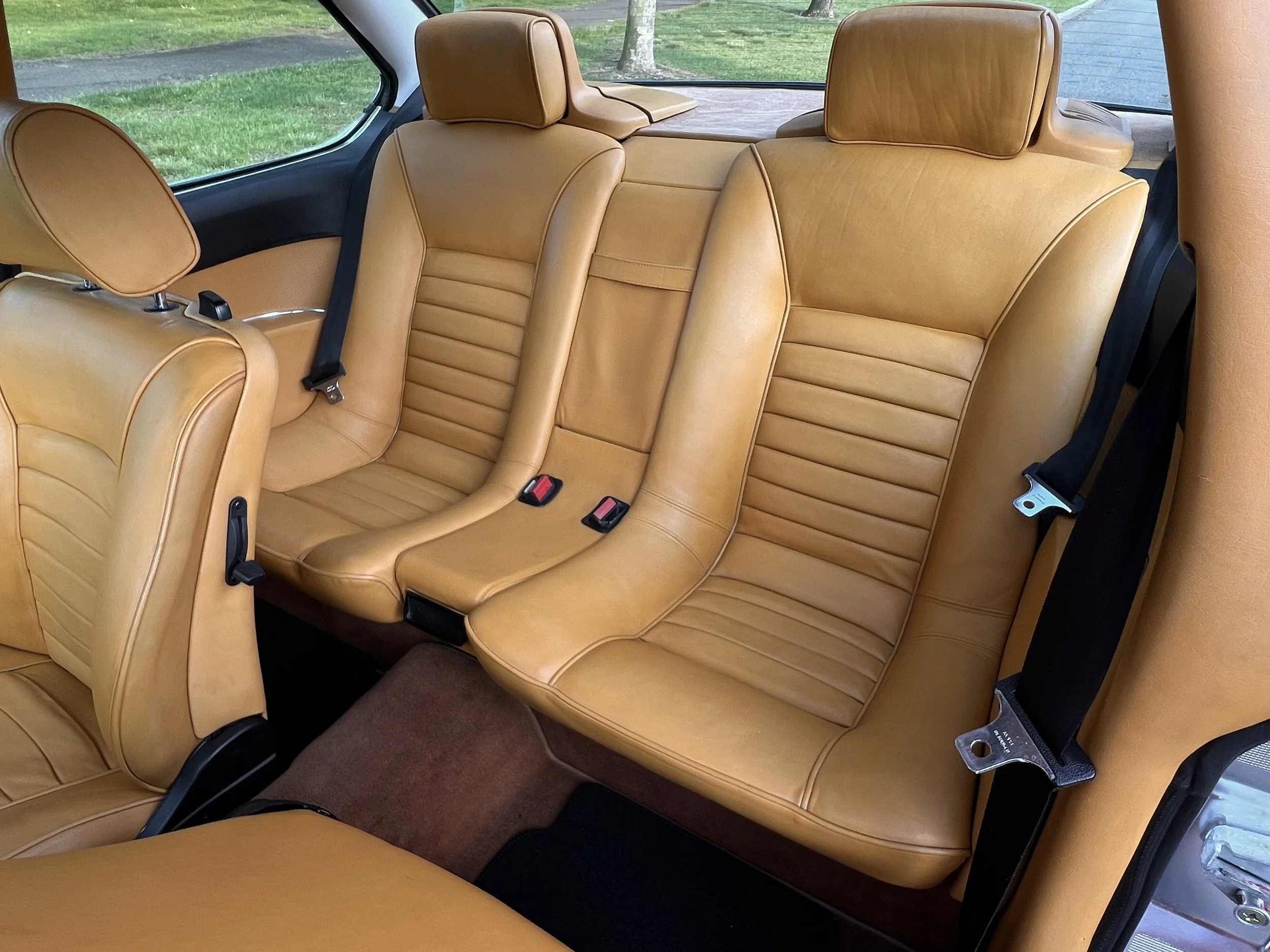 The interior of a 1977 BMW 630 csi car showing tan leather seats, two in the back row with headrests and seatbelts, and a part of the front seat.