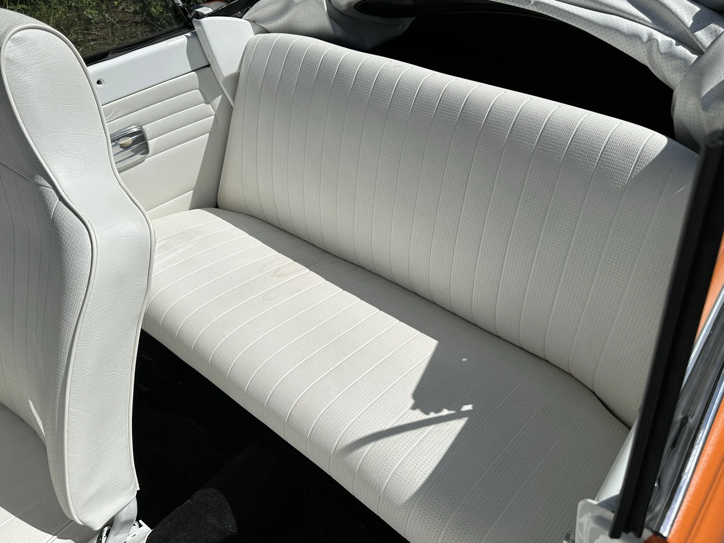 The interior of a vintage car showing a white leather bench seat and part of a front seat, with a black floor and a window.