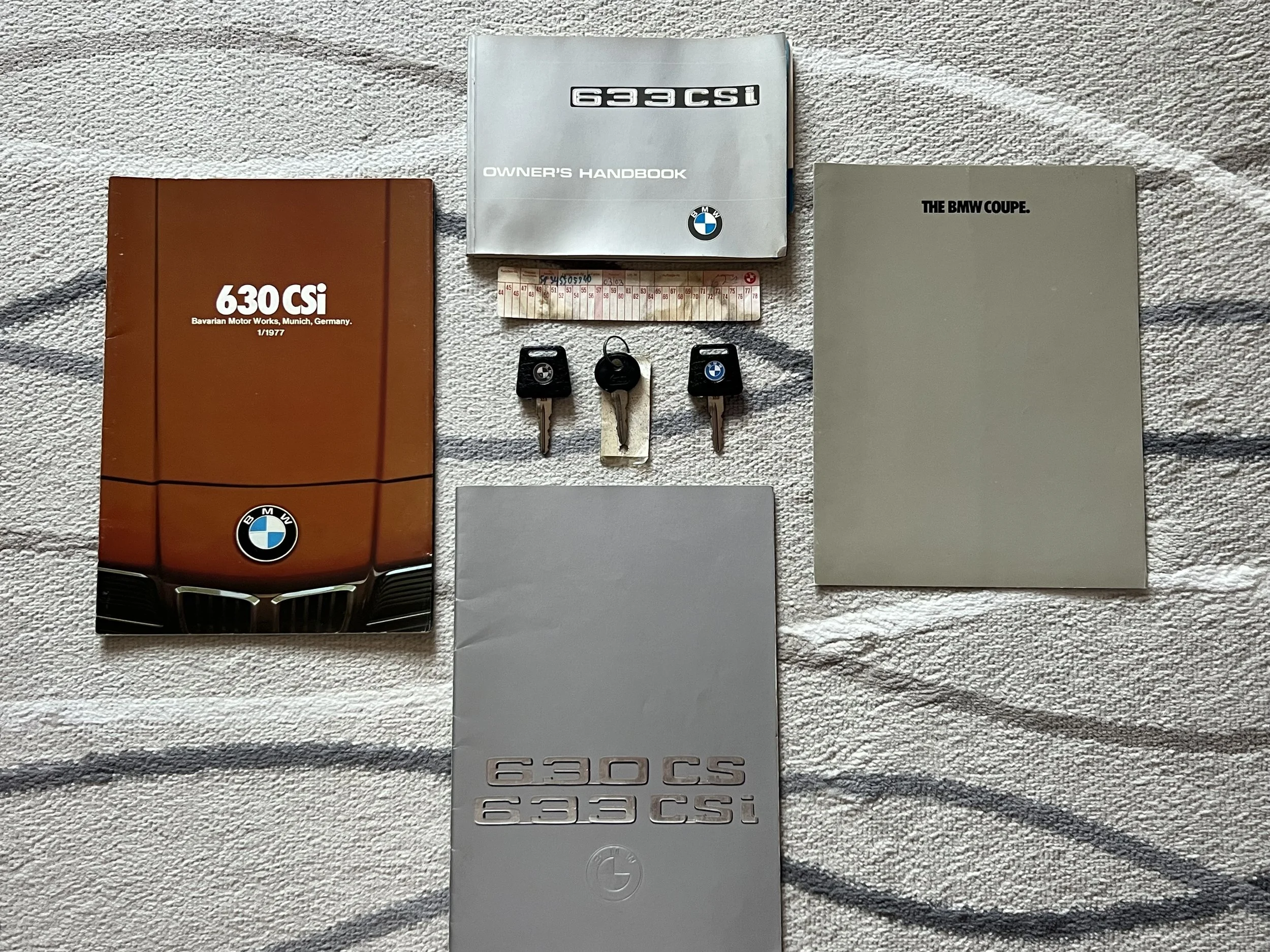 Collection of 1977 BMW 630 csi car manuals, keys, and accessories laid out on a light-colored fabric surface. The items include three car keys, several manuals for BMW models 630 CSI and 633 CSI, a ruler, and a white manual.